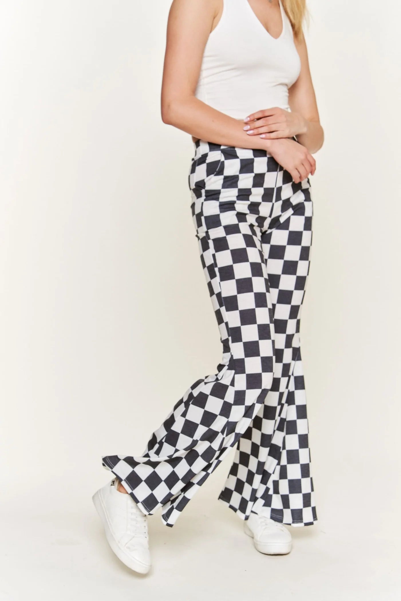 Woman wearing black and white checkered flared pants paired with white tank top and white sneakers against plain backdrop