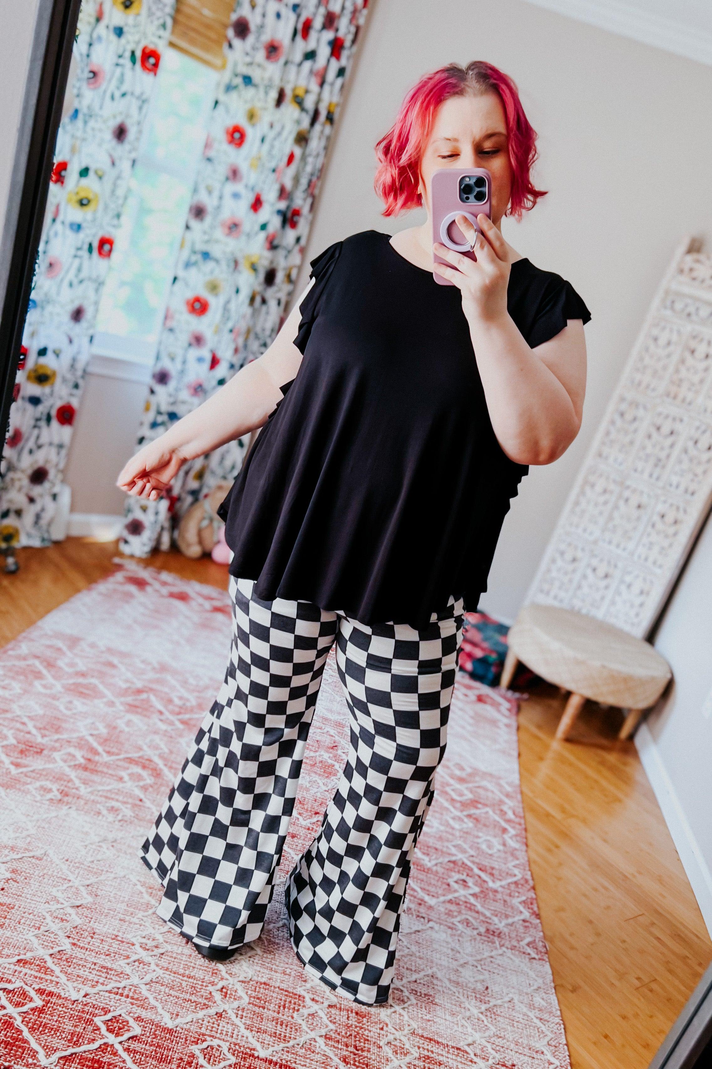 Plus-size woman in black top and black and white checkered flared pants taking mirror selfie indoors