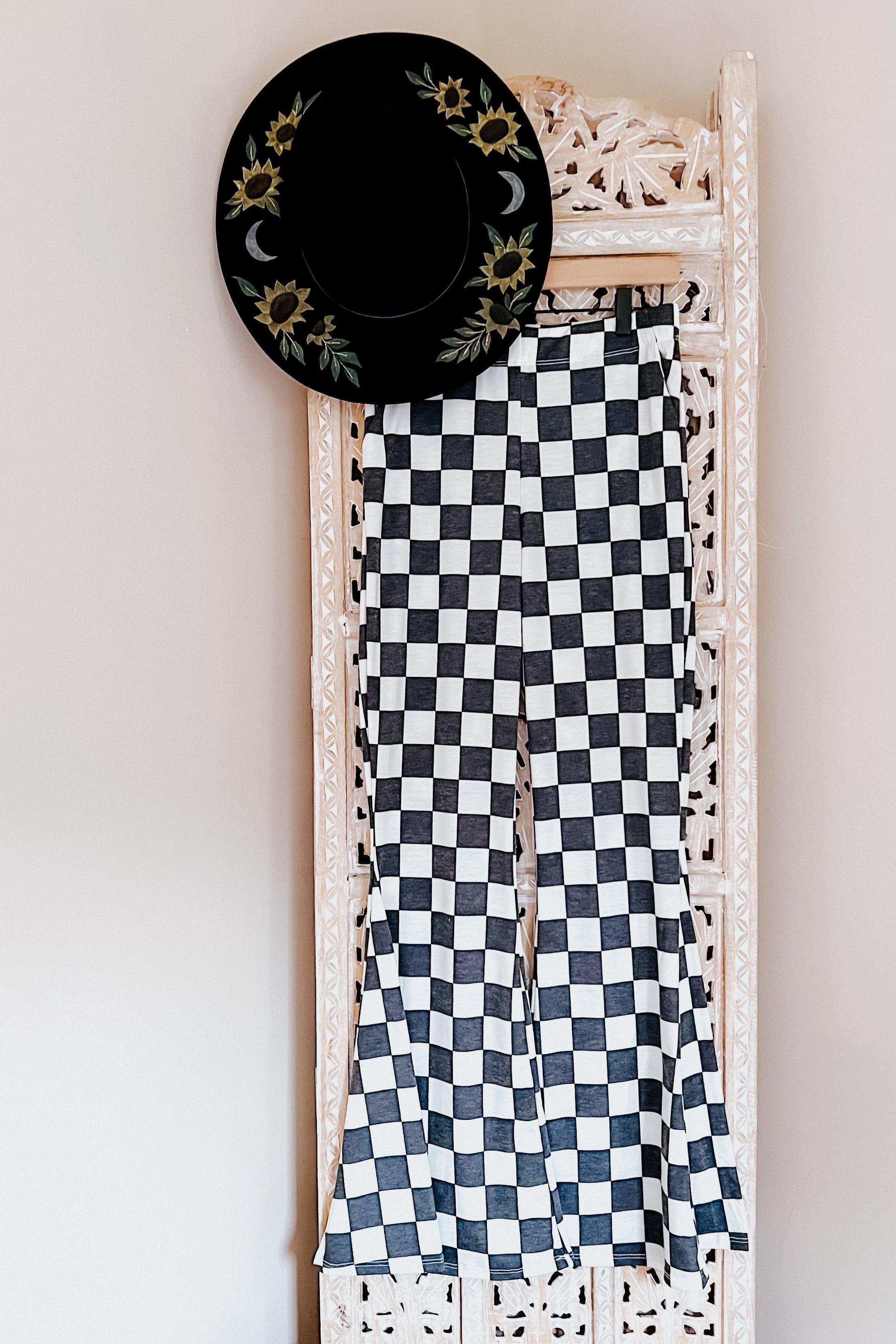 Black and white checkered flared pants hanging on decorative white carved screen with black hat featuring sunflower embroidery