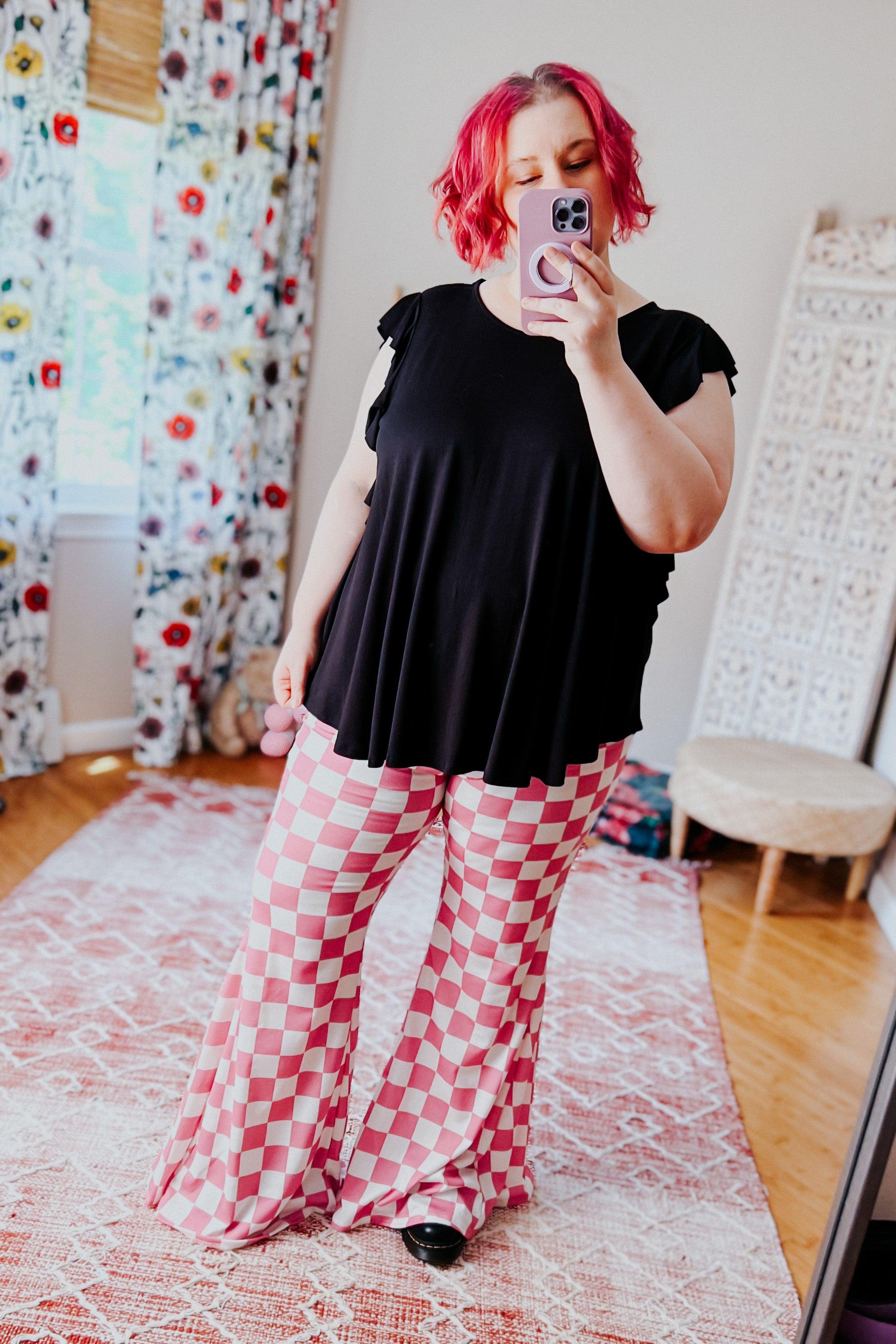 Woman taking mirror selfie wearing pink and white checkered flared pants with black top in bright room