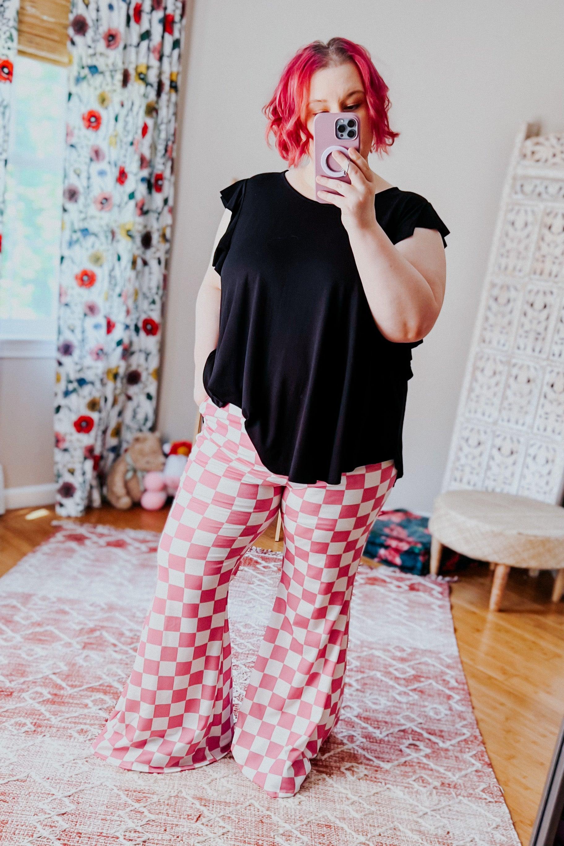 Woman with pink hair wearing pink and white checkered flared pants and black top taking a mirror selfie indoors