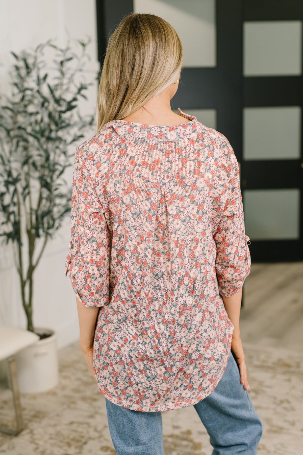 Back view of a woman wearing a peach floral half sleeve top with a rounded hem and blue jeans in a bright home interior