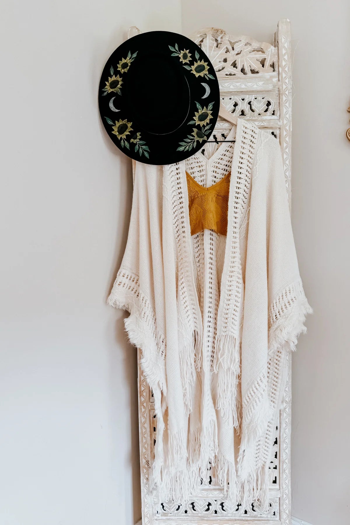Cream bohemian ruana with open knit details and fringe hanging on carved white wooden screen with black floral hat