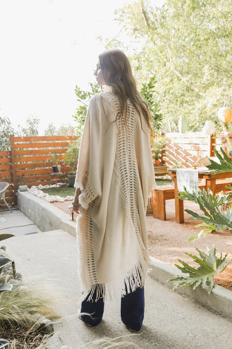 Woman wearing a cream ruana with textured knit center panel and fringe, standing outside near wooden fence and plants