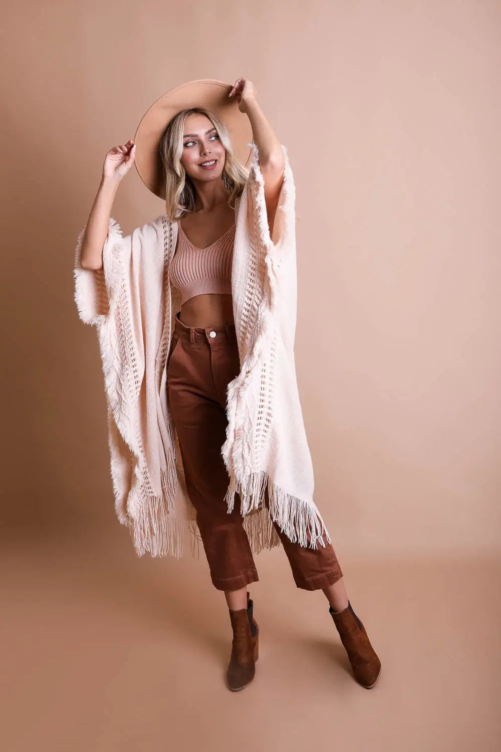 Blonde woman wearing cream frayed border bohemian ruana with wide sleeves, styled with cropped knit top, rust pants, and tan hat