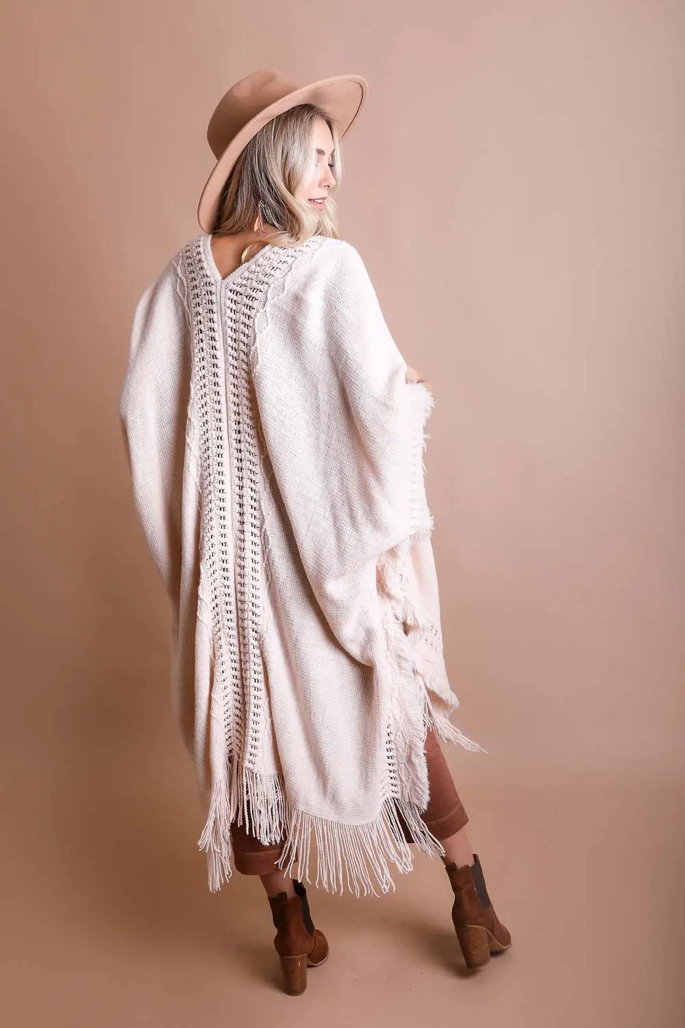 Woman in tan hat wearing a cream boho ruana with open knit details and long fringe standing against beige background