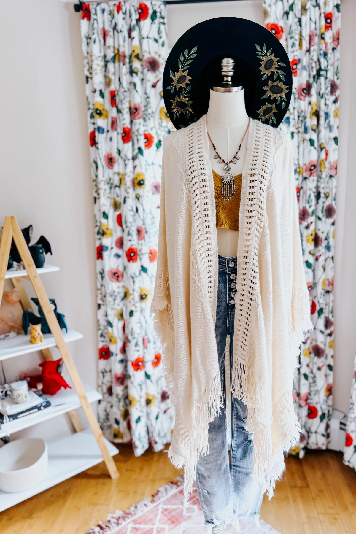 Cream bohemian ruana with frayed edges layered over mustard knit top and high-waisted light wash jeans on mannequin