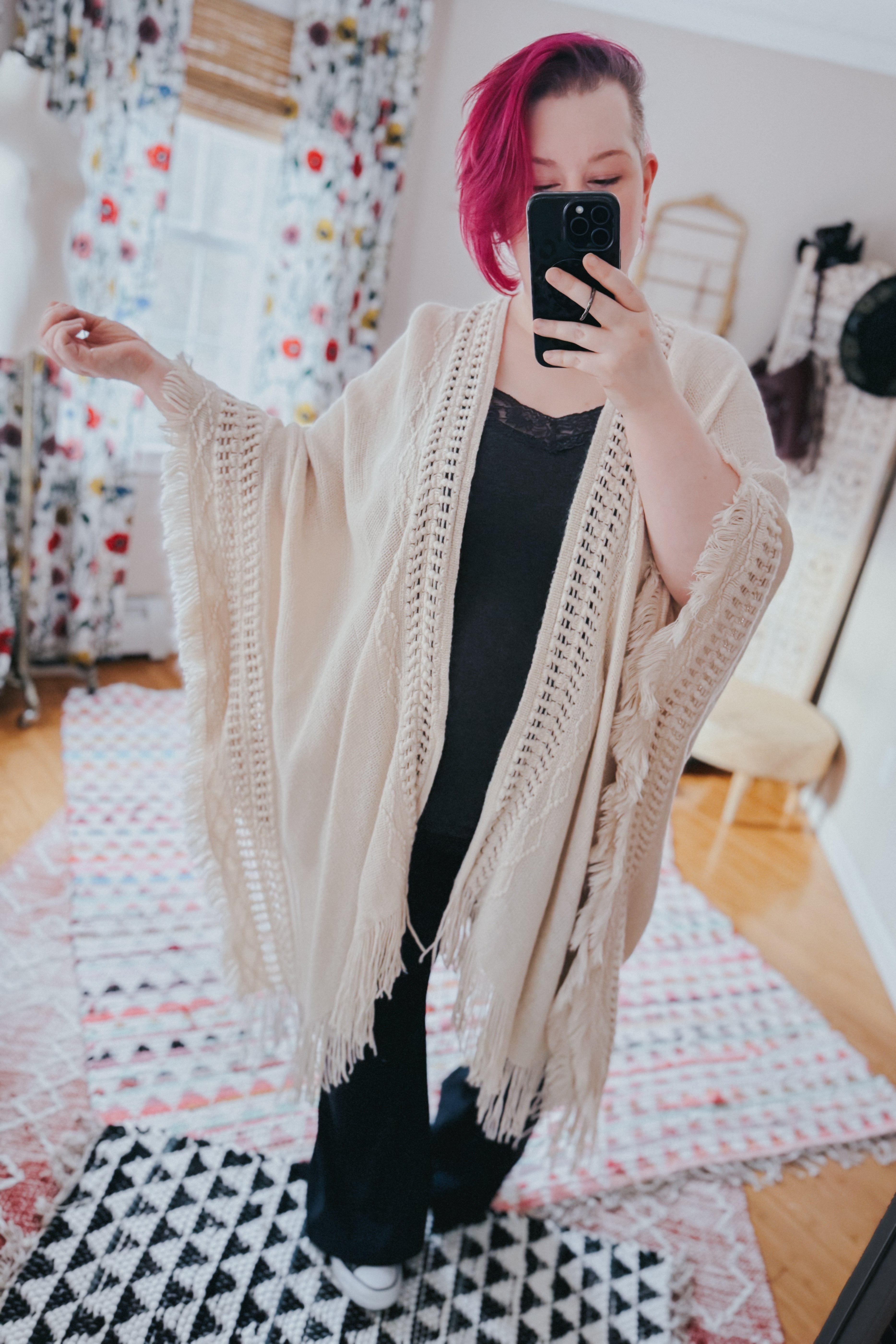 Woman with short magenta hair wearing a cream ruana with frayed edges over black outfit taking a mirror selfie indoors