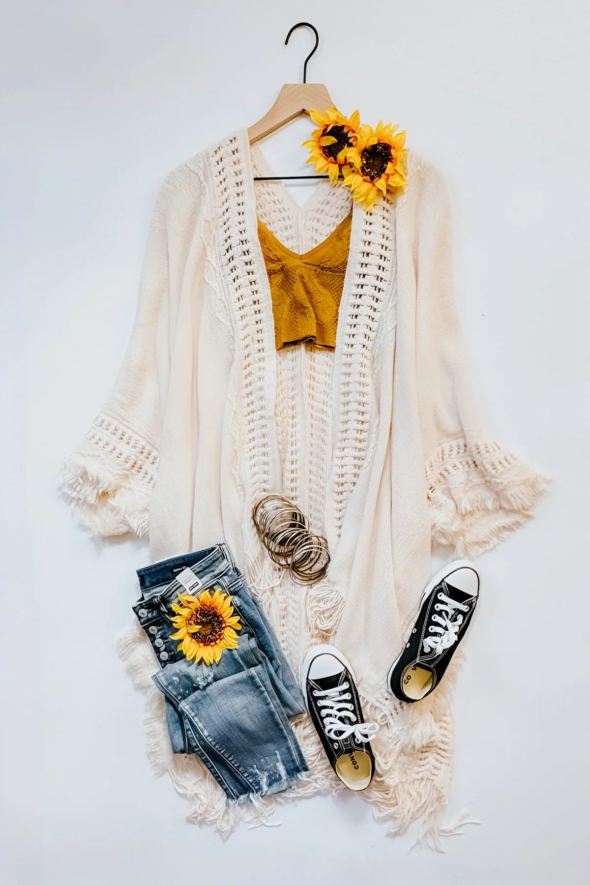 Cream bohemian ruana with frayed sleeves styled with mustard crop top, distressed jeans, black sneakers, and sunflowers on white background