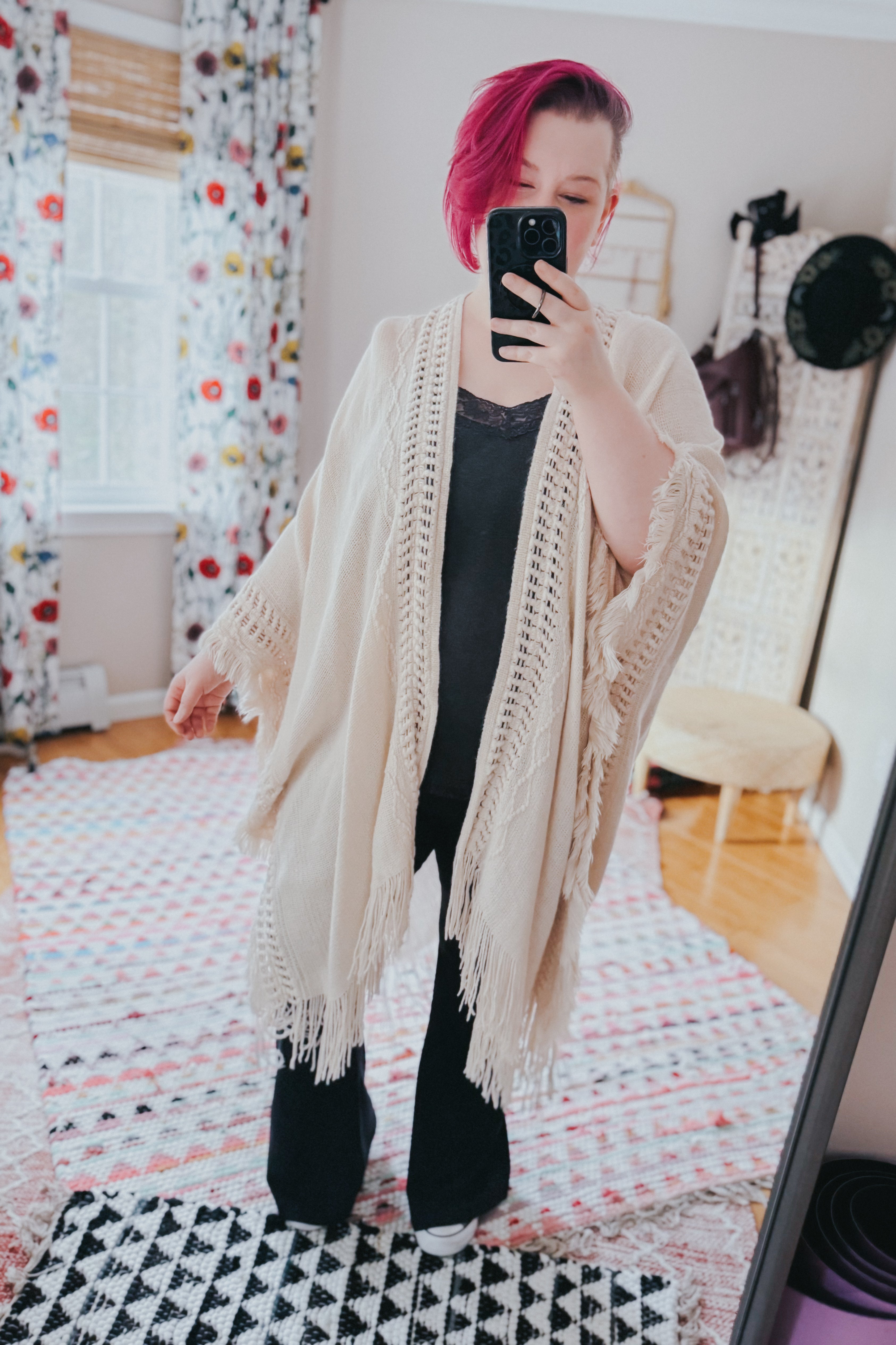 Woman with short magenta hair wearing a cream frayed border ruana over black outfit taking mirror selfie indoors