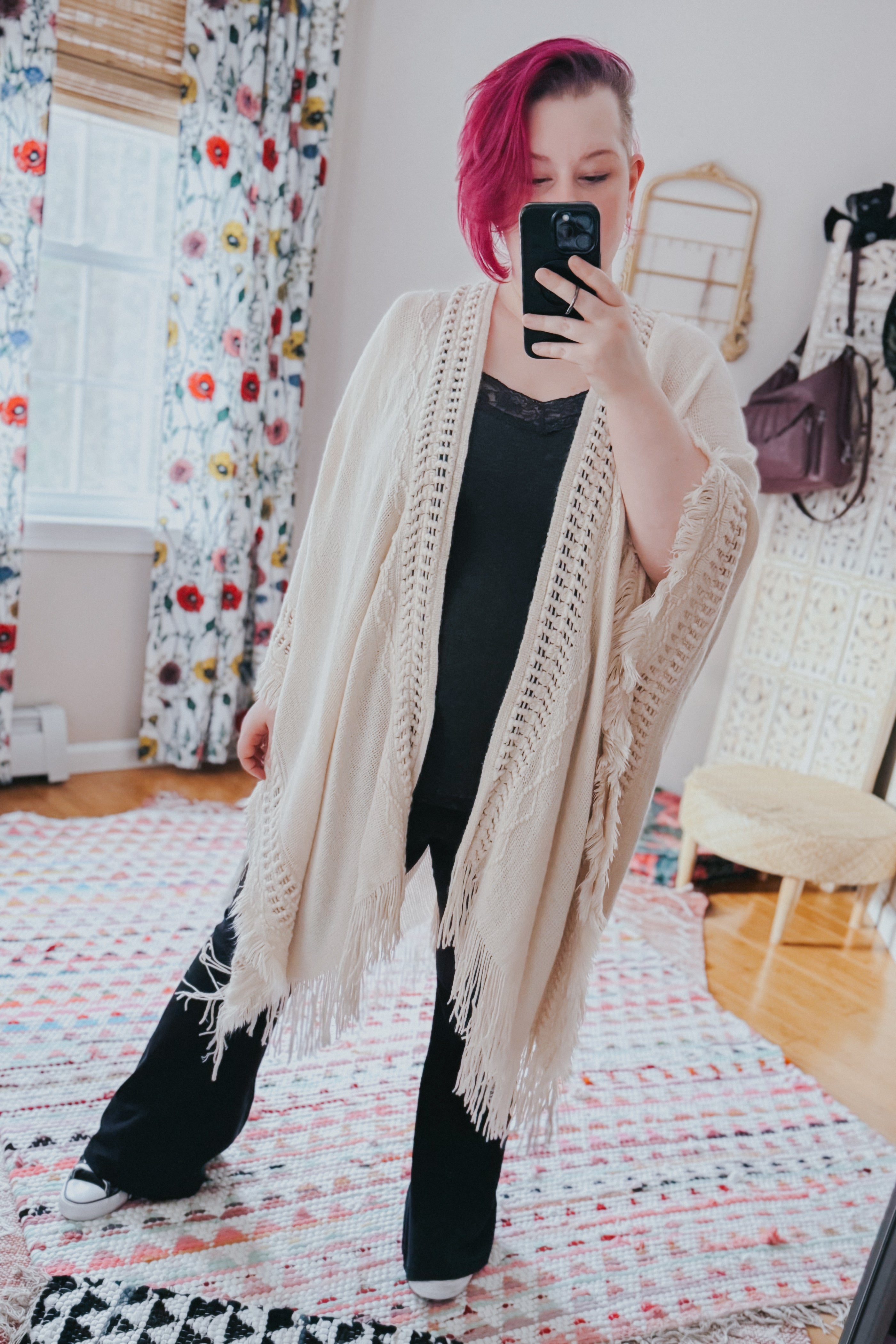 Woman with short pink hair wearing a cream crochet ruana with fringed edges over black outfit taking a mirror selfie