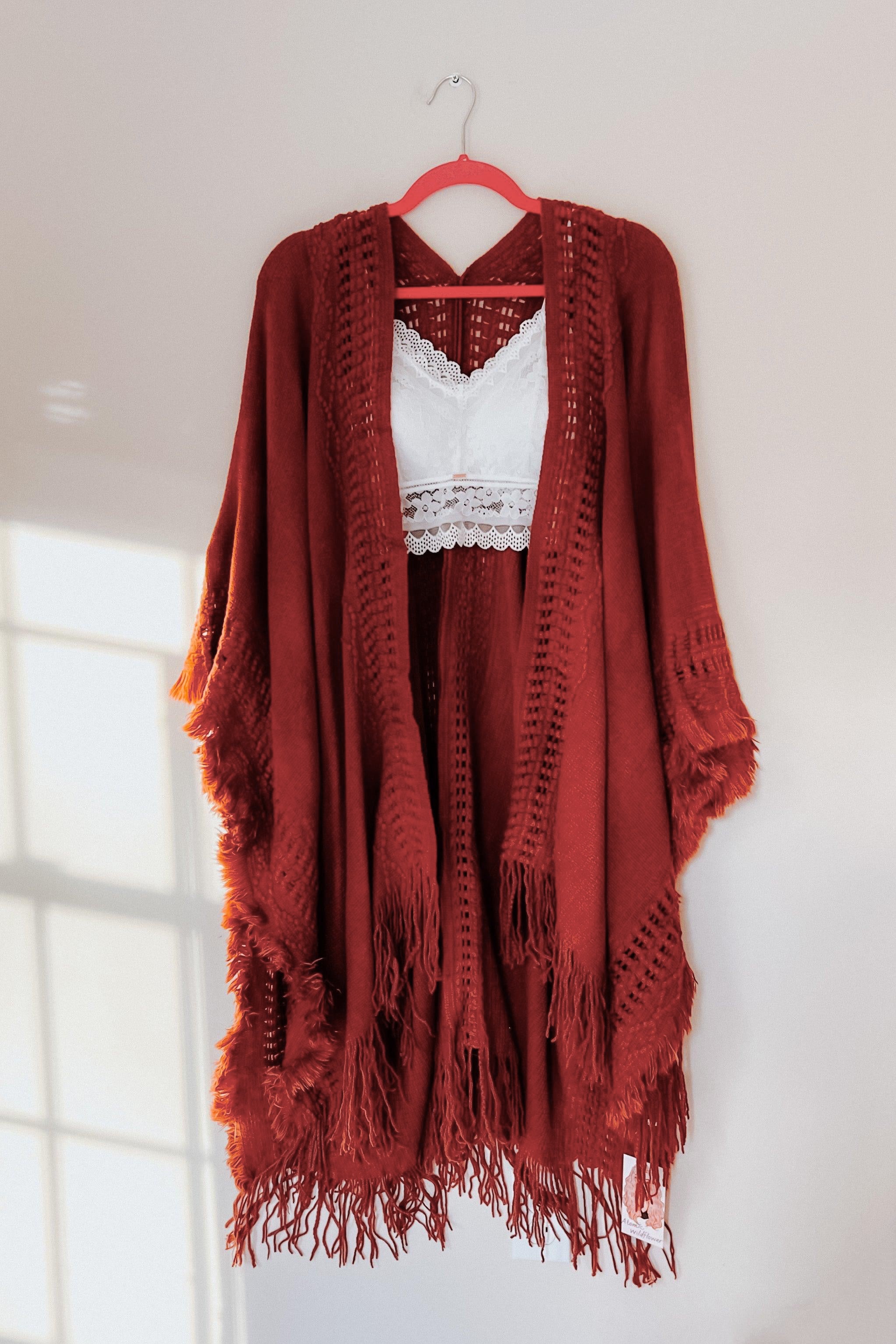 Rust-colored bohemian ruana with frayed edges layered over a white lace bralette hanging on a pink hanger