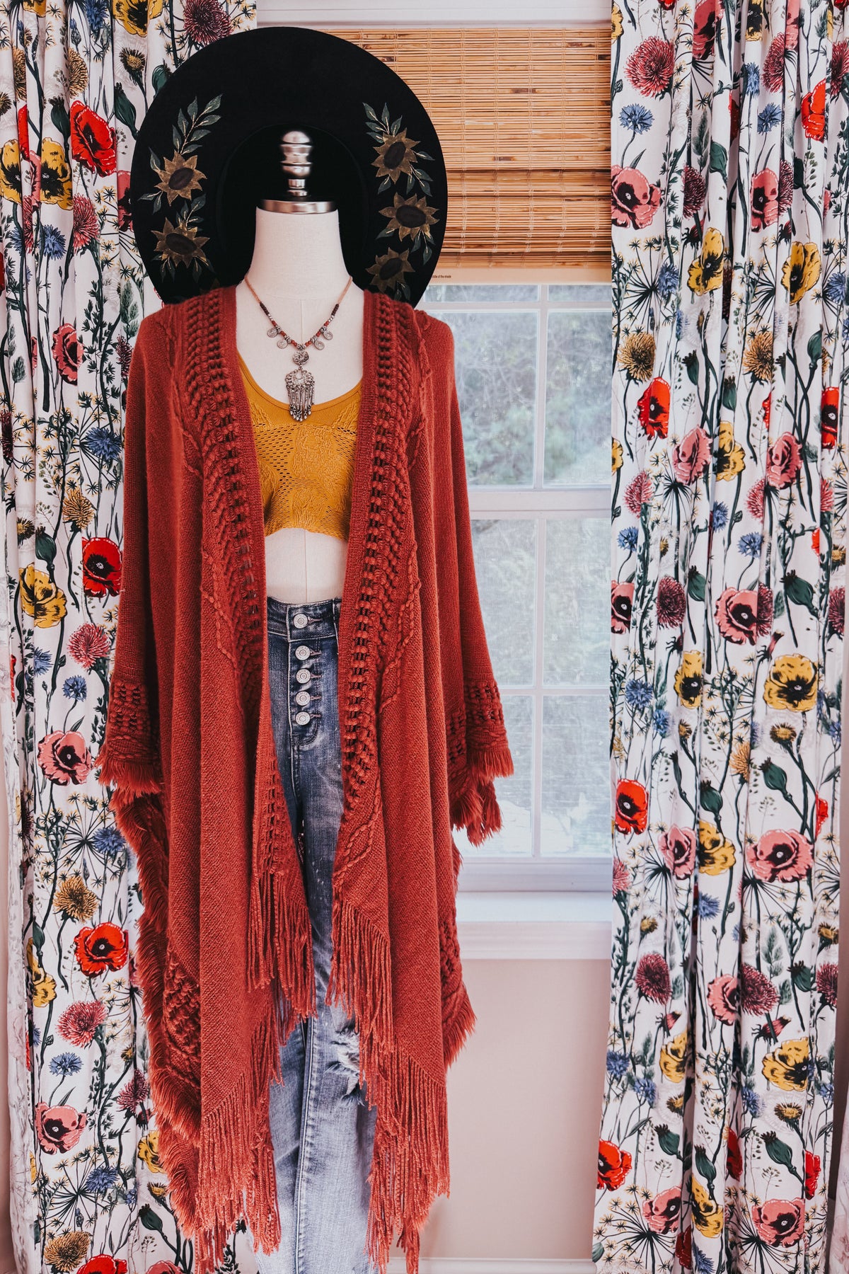 Mannequin wearing a rust bohemian ruana with open front long fringe mustard top distressed jeans and layered necklaces indoors