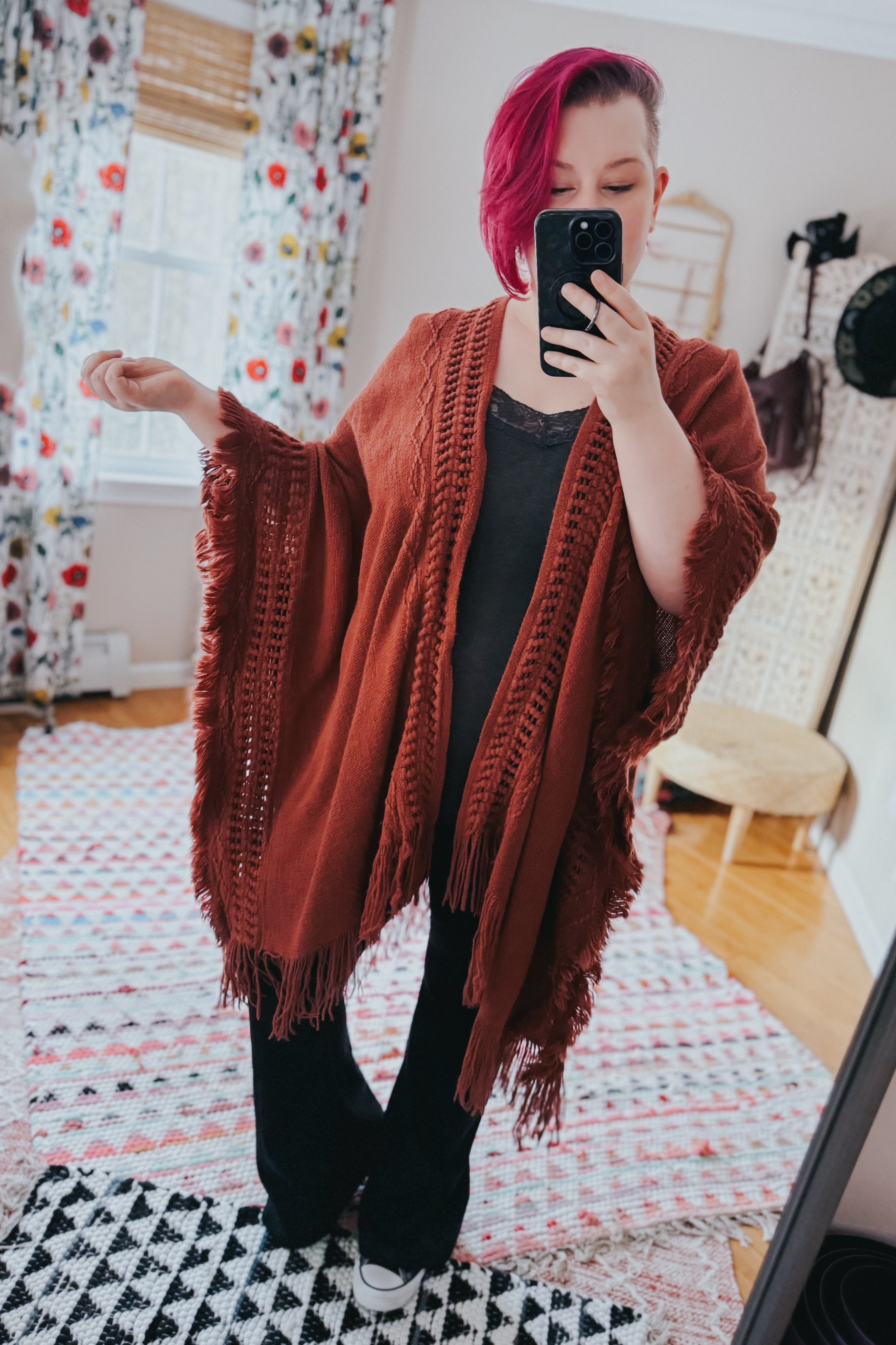 Woman with magenta hair wearing rust-colored fringed bohemian ruana over black outfit indoors taking mirror selfie
