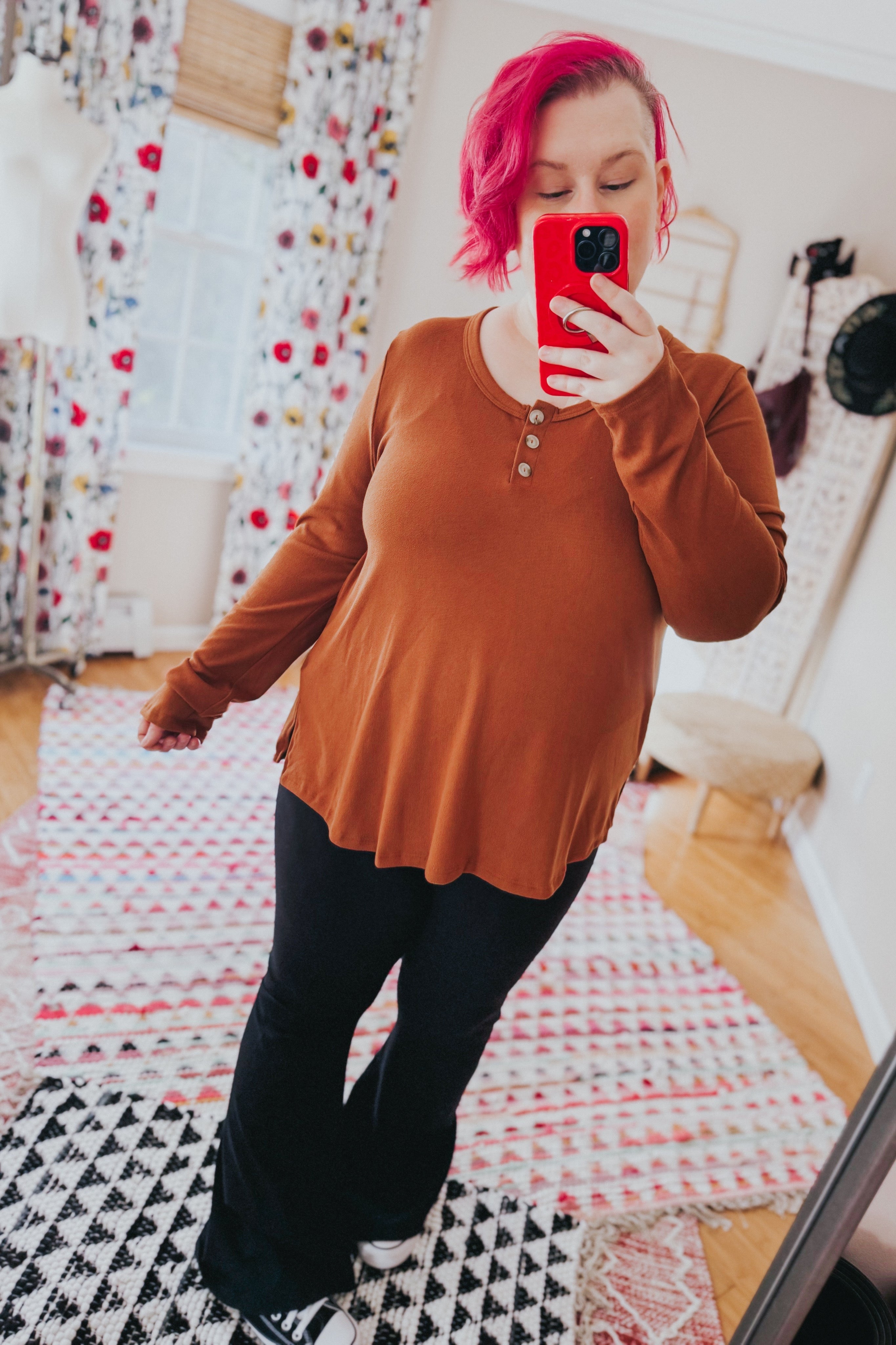Woman taking a mirror selfie wearing a rust scoop neck top with button details and black pants in a bright room