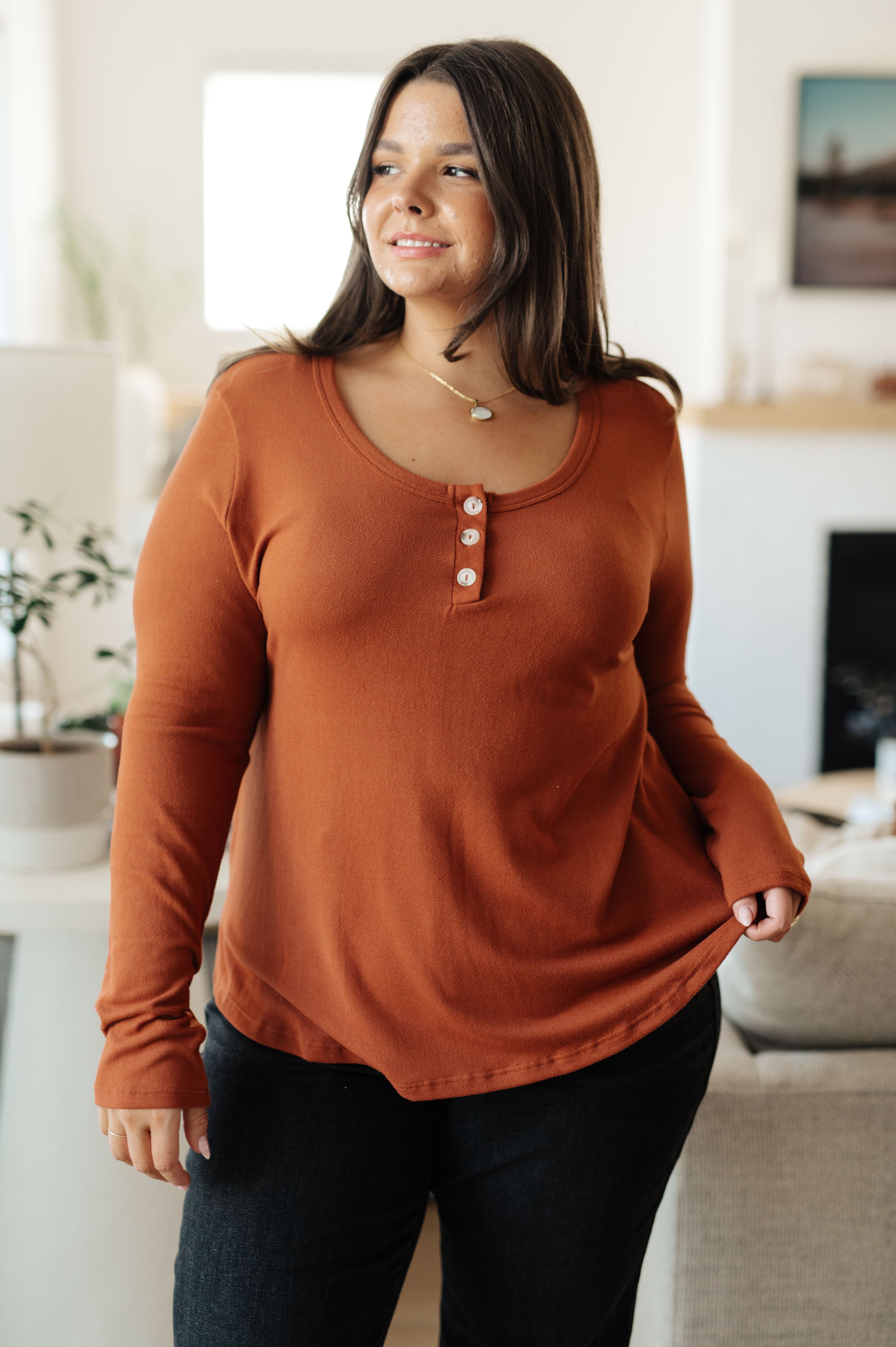 Plus size woman smiling and wearing a rust scoop neck long sleeve top with three buttons in a casual home setting