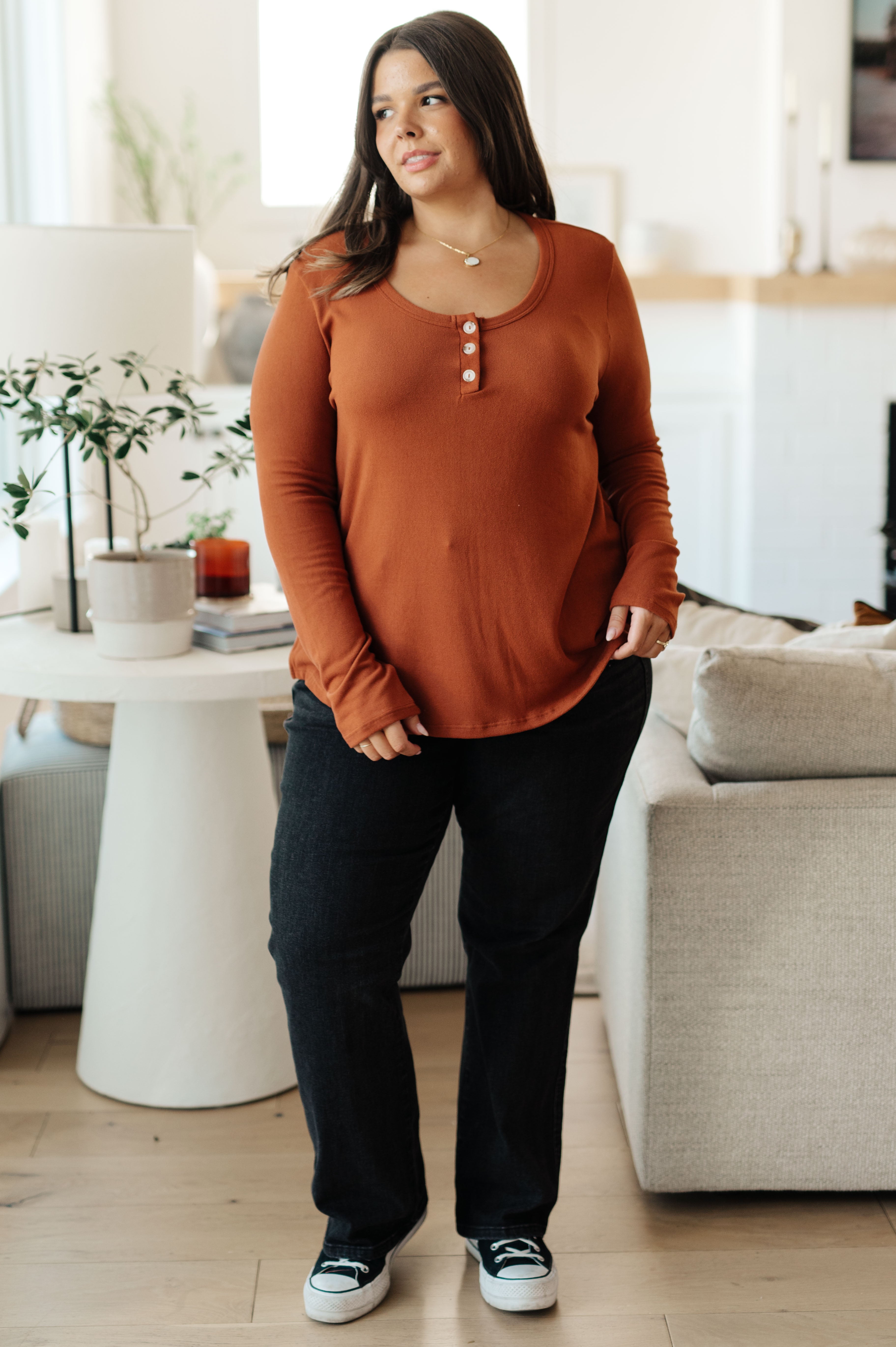 Plus size woman wearing a rust scoop neck top with button placket styled with black pants and sneakers in a cozy living room