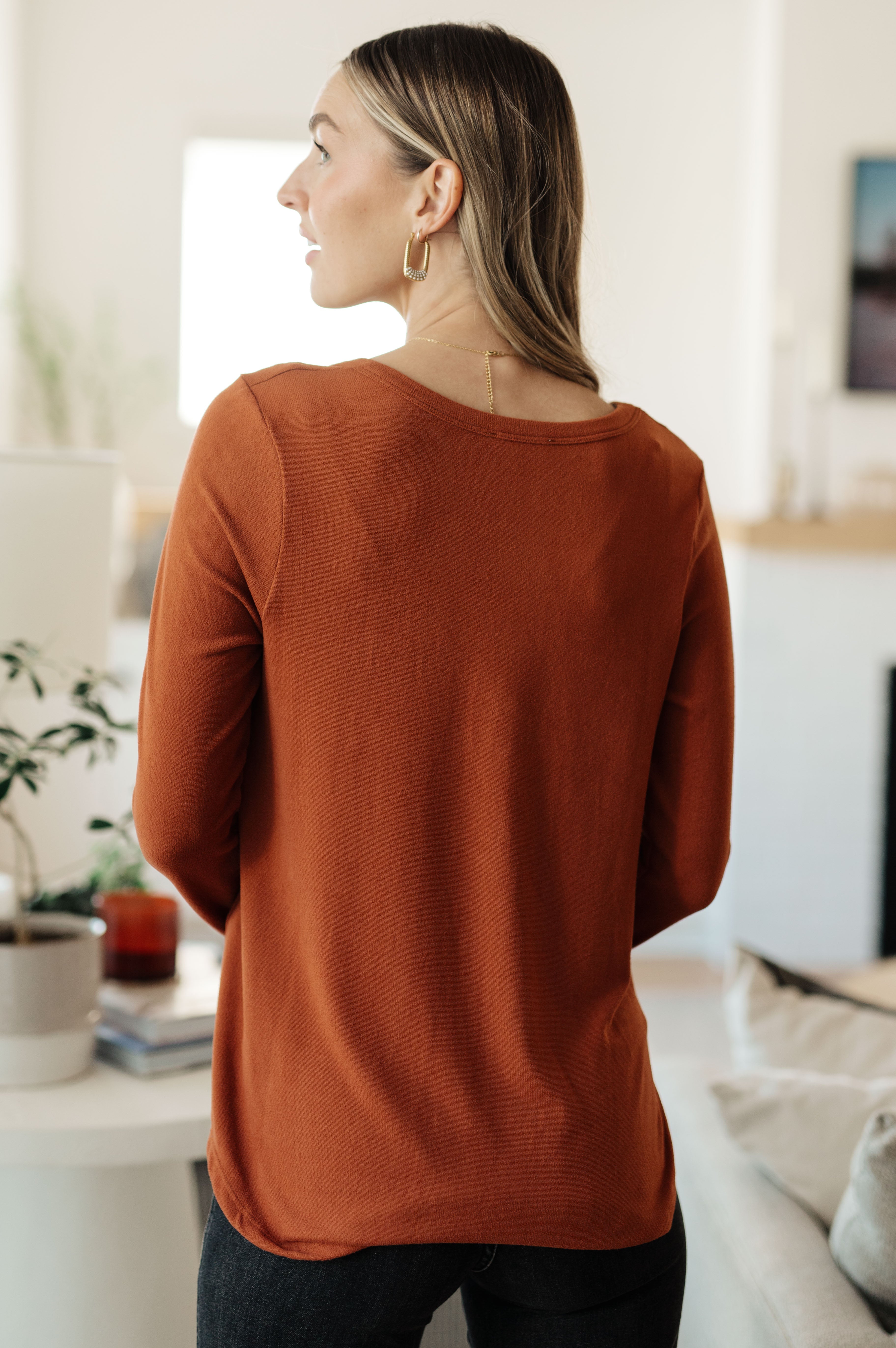 Woman seen from behind wearing a rust-colored scoop neck long sleeve top in a cozy indoor setting