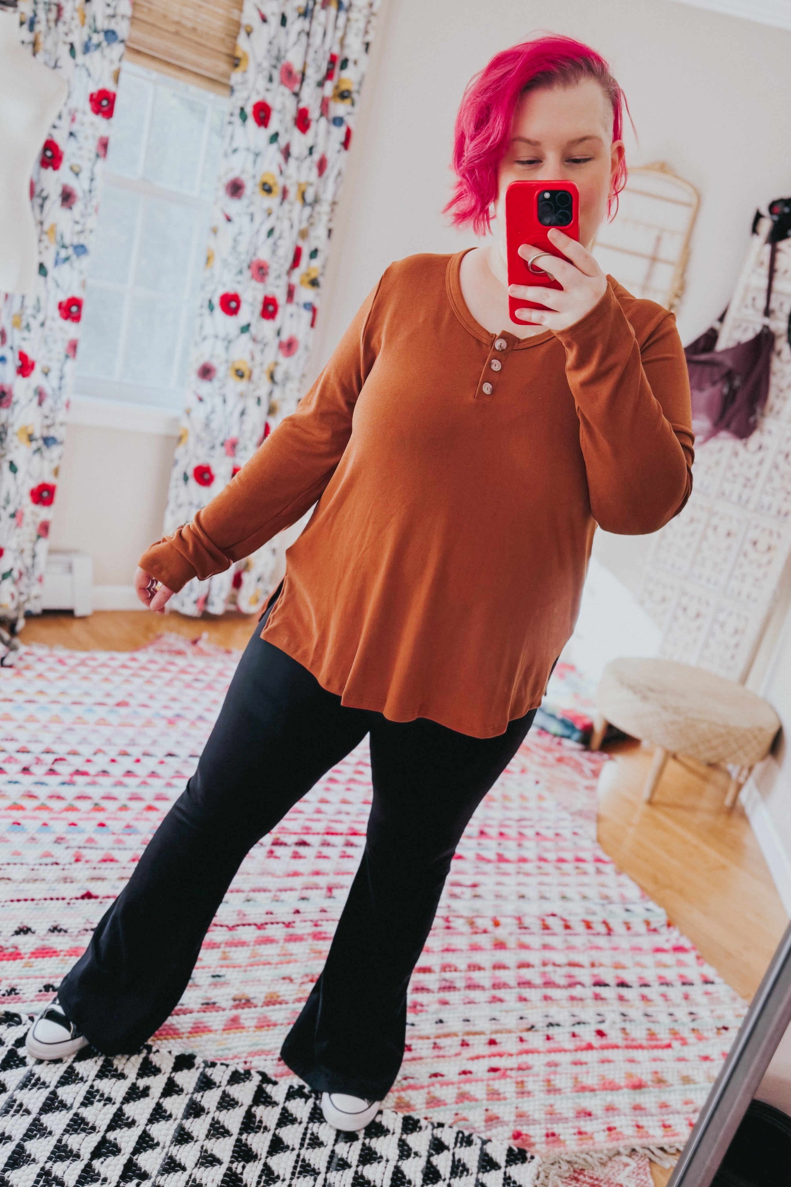 Woman taking a mirror selfie wearing a rust scoop neck top with button accents and black pants in colorful room