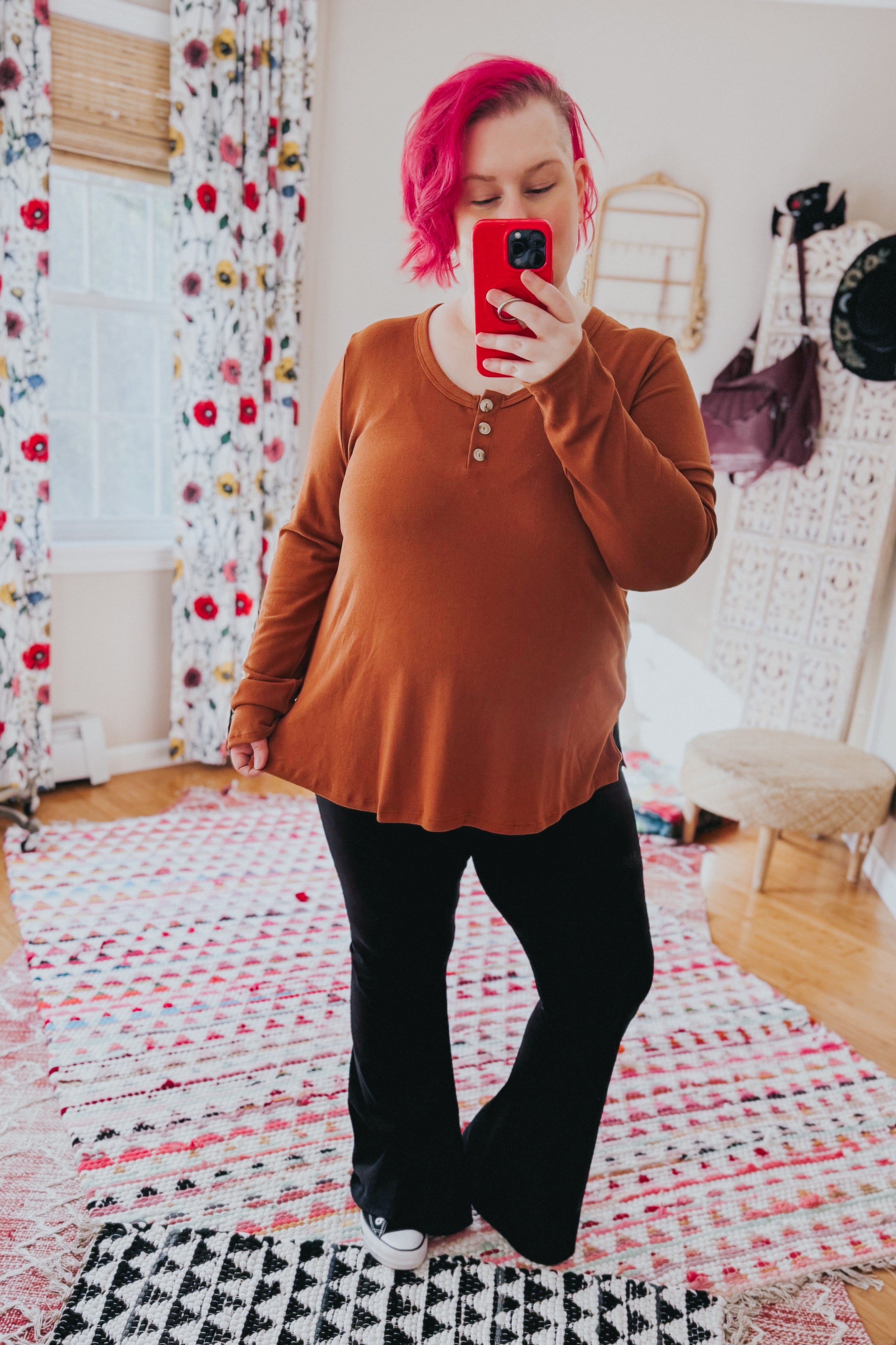Woman wearing a rust scoop neck top with button details paired with black pants taking a mirror selfie indoors