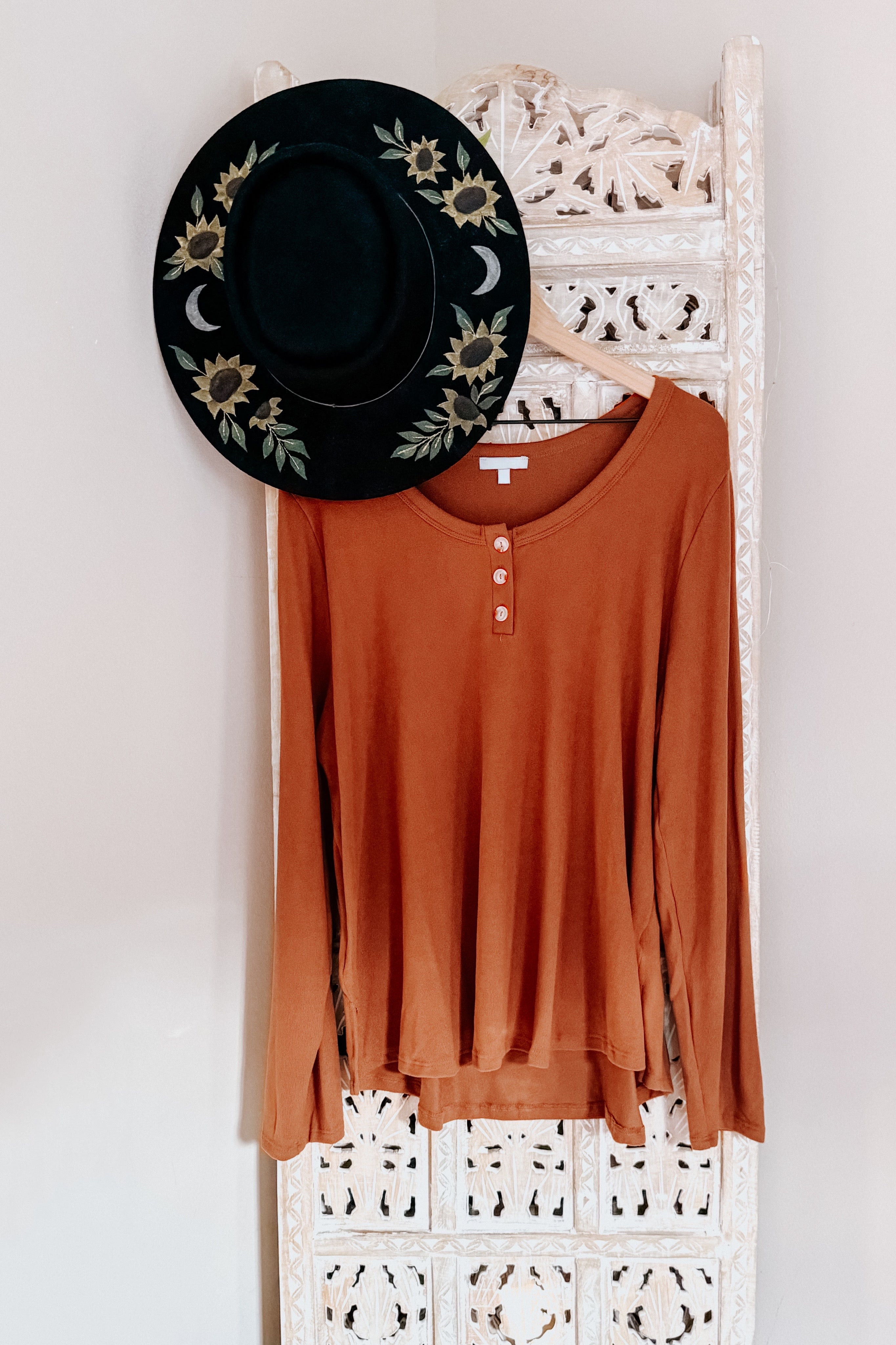 Rust-colored scoop neck long sleeve top with three-button placket hanging on carved white wooden panel with black hat