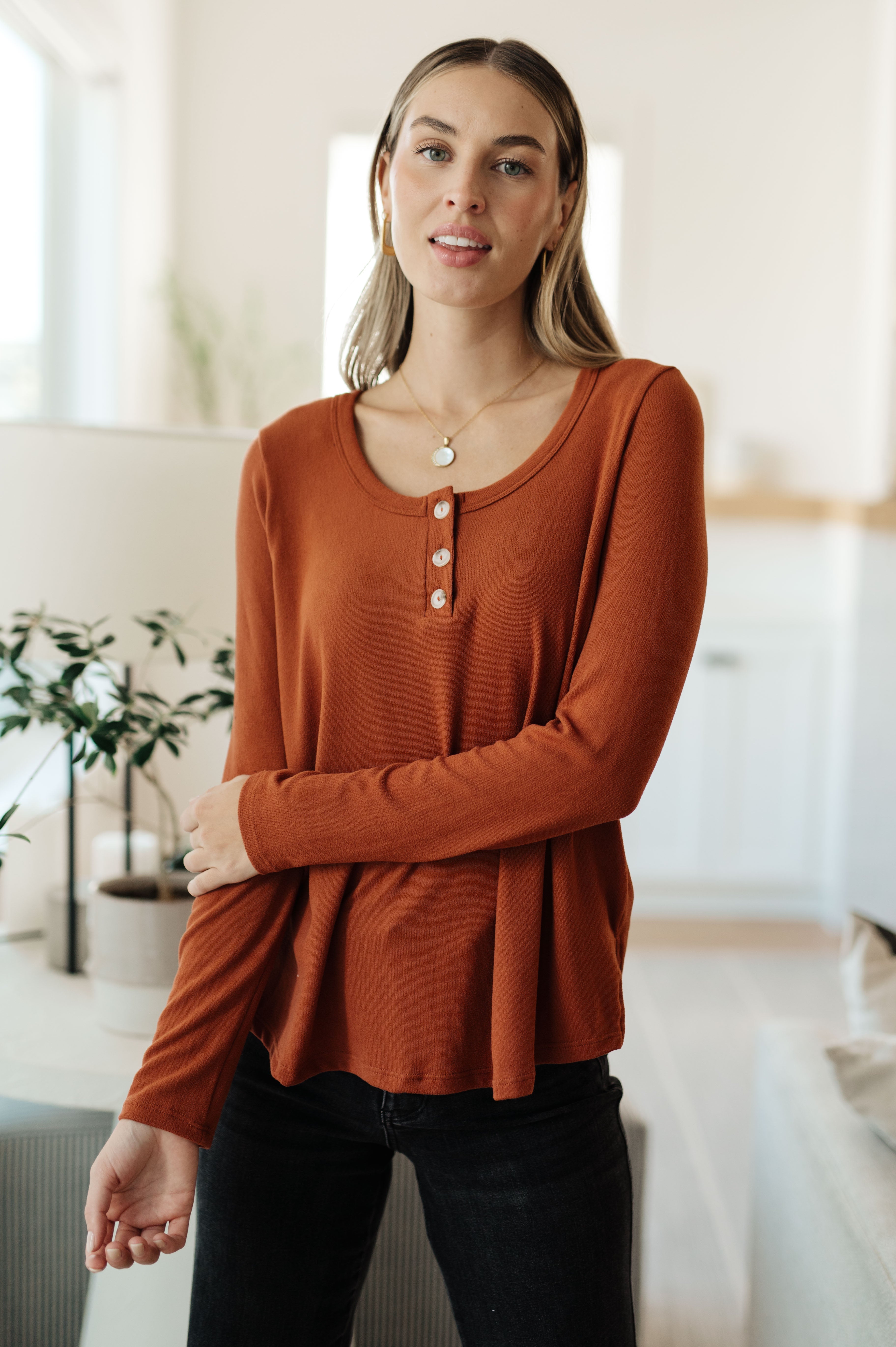 Woman wearing a burnt orange scoop neck top with button placket styled casually indoors with long sleeves and black jeans