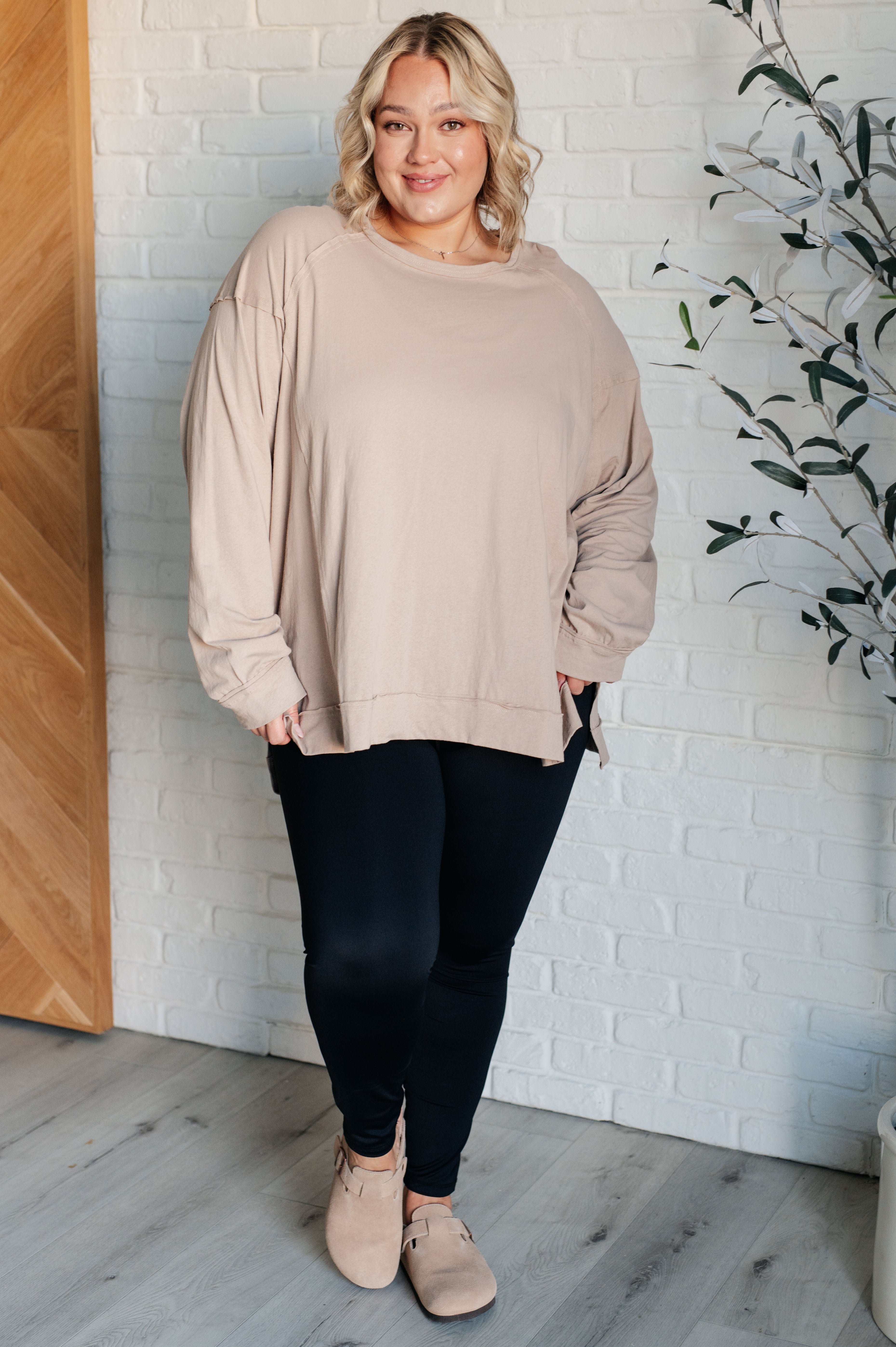 Smiling woman wearing a loose beige long sleeve top and black leggings standing indoors near white brick wall and plant