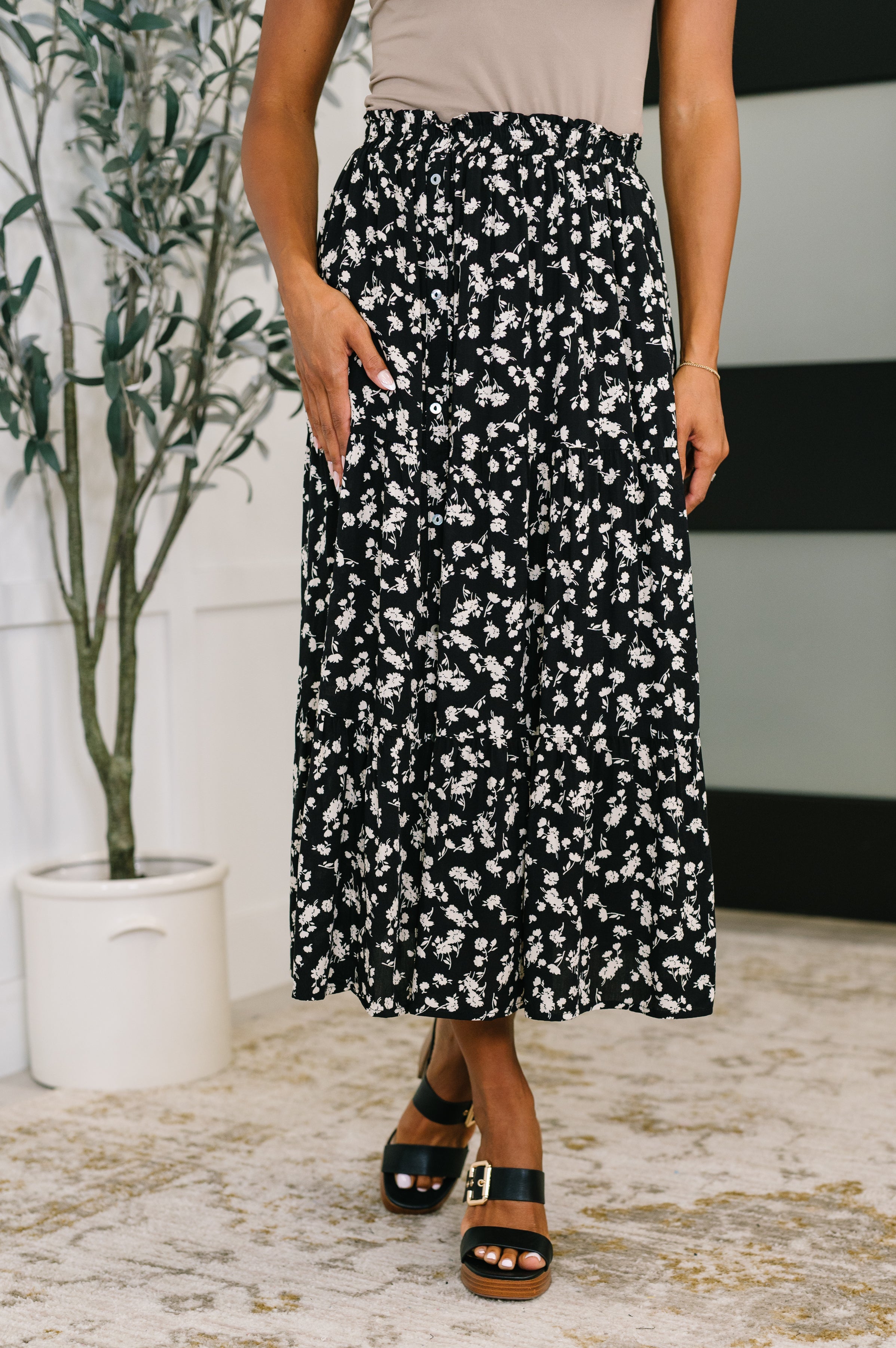 Woman wearing a black floral skirt with side pockets paired with black platform sandals indoors by potted plant