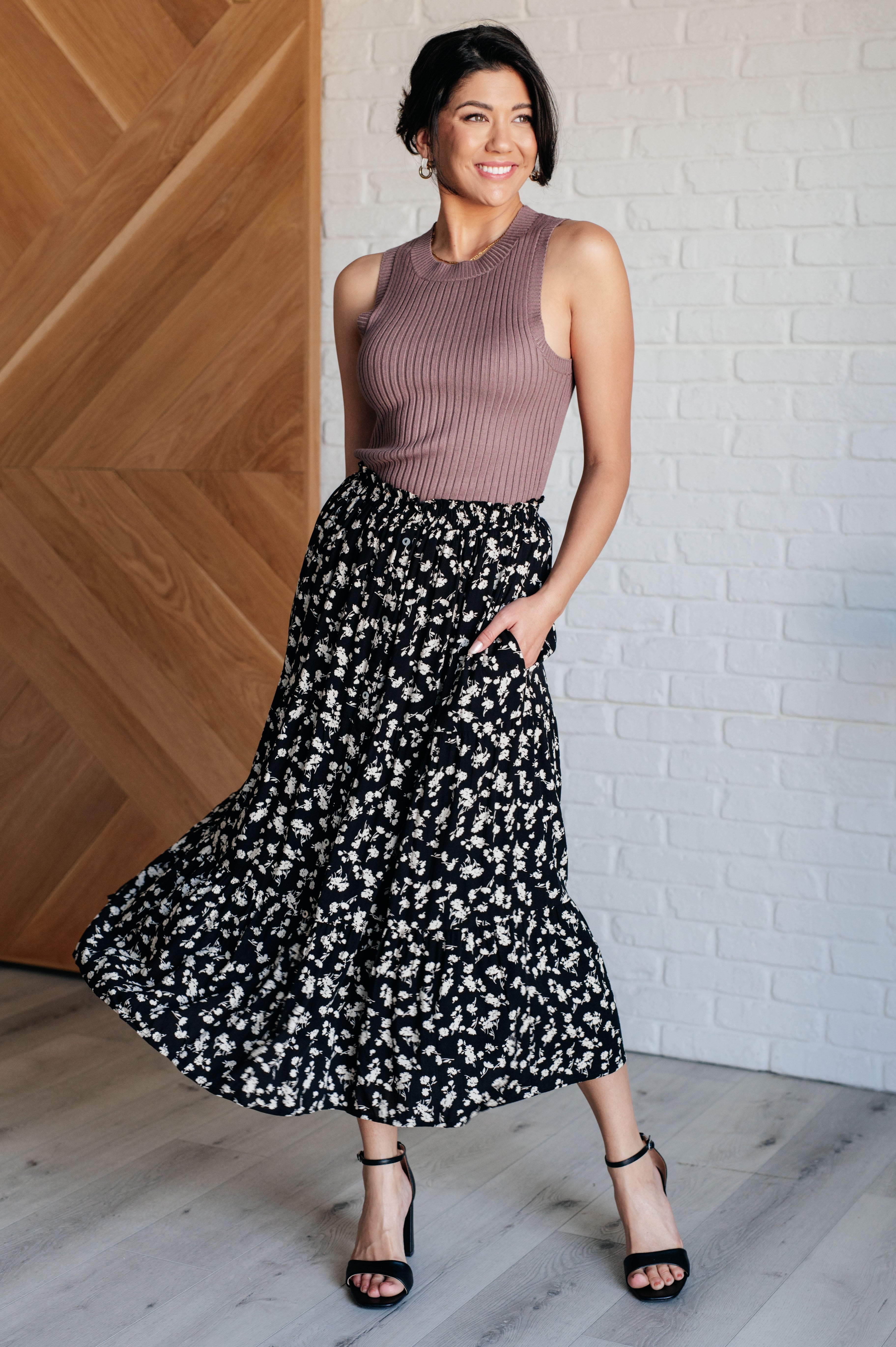 Woman wearing a black floral skirt with side pockets paired with a sleeveless ribbed mauve top and black heels indoors