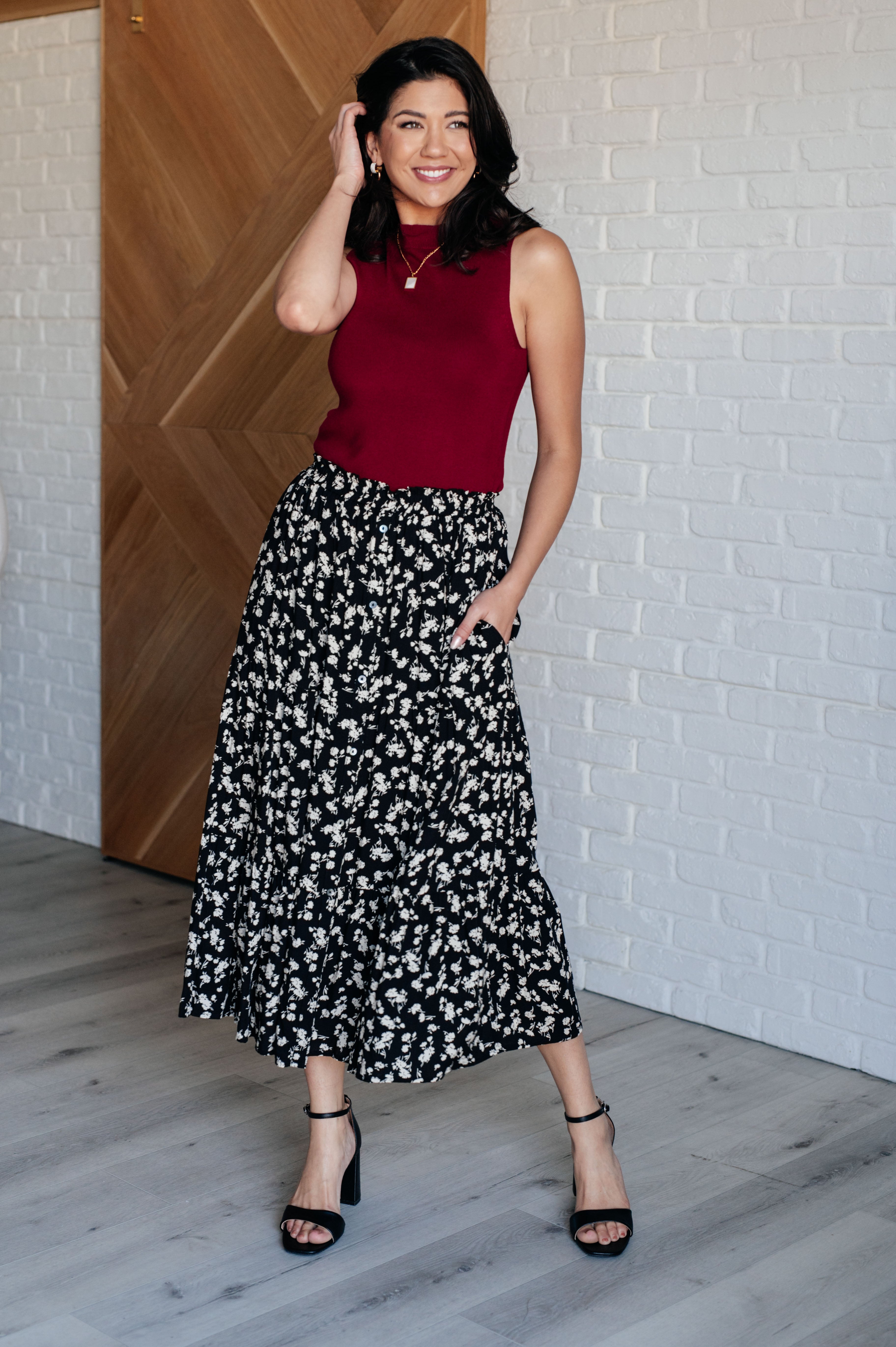 Smiling woman wearing a black floral skirt with side pockets and black ankle-strap heels standing indoors