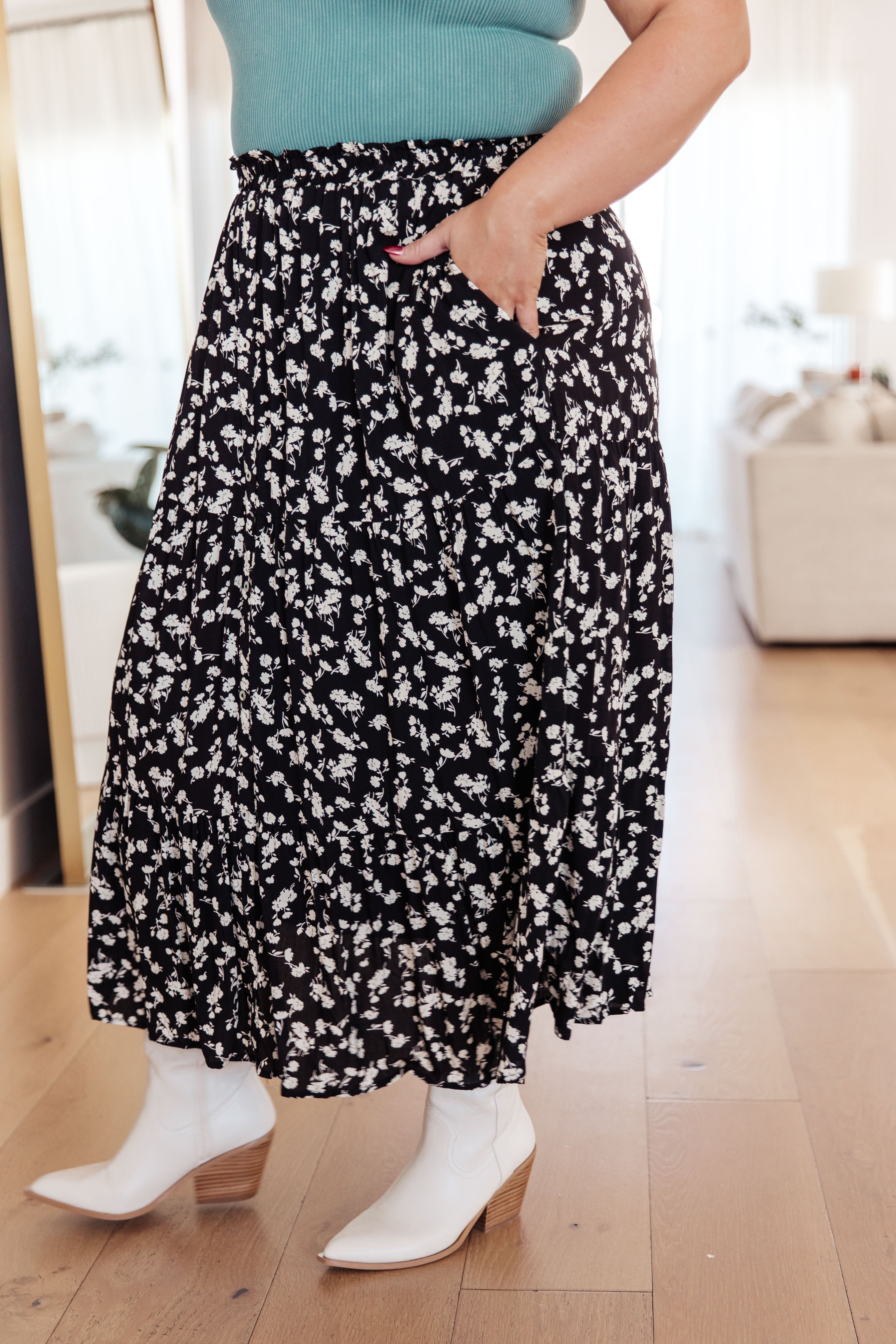Woman with hand in side pocket of black floral midi skirt paired with white ankle boots on hardwood floor