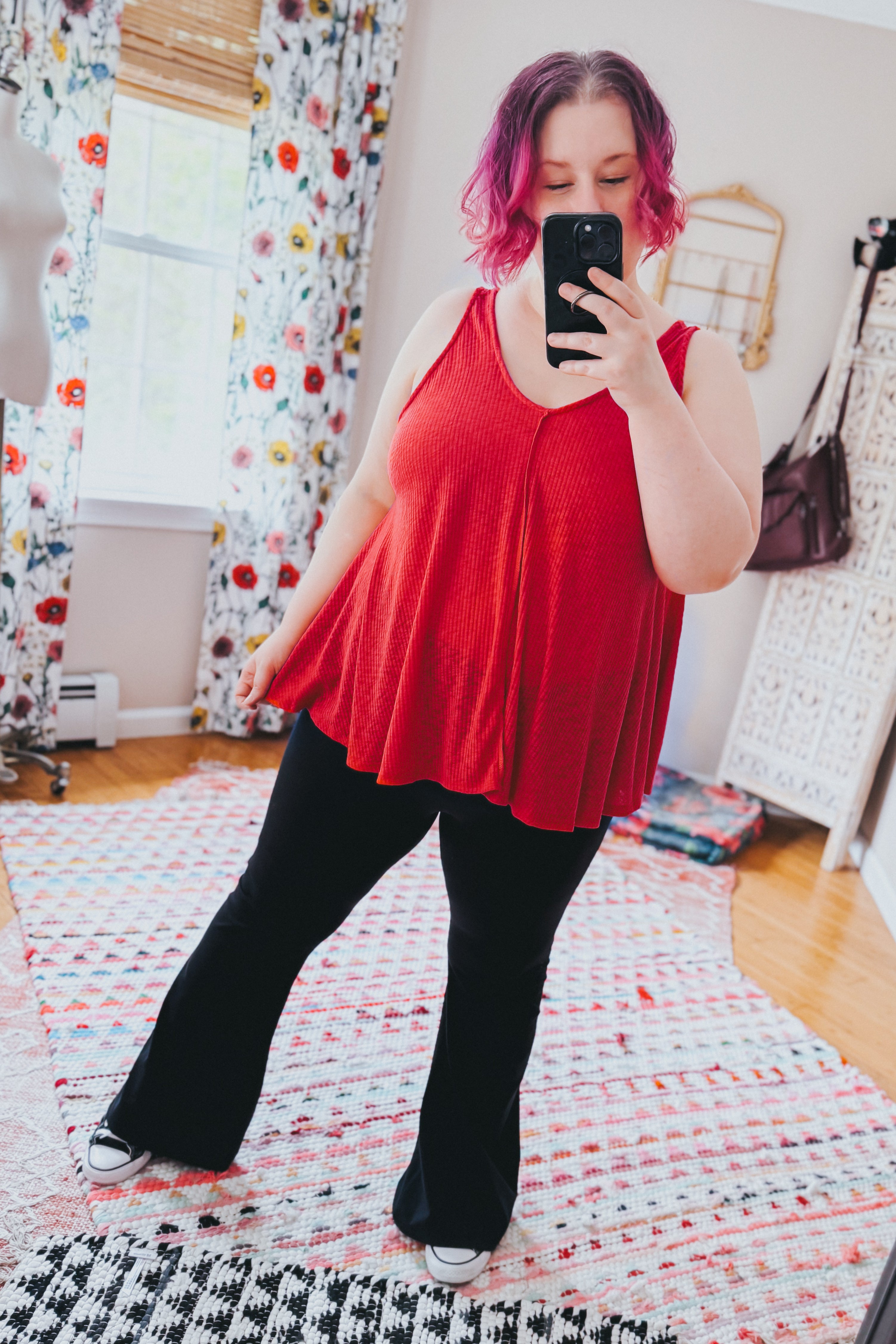 Woman with short pink hair wearing a loose flowing red sleeveless tank top and black flared pants taking indoor mirror selfie