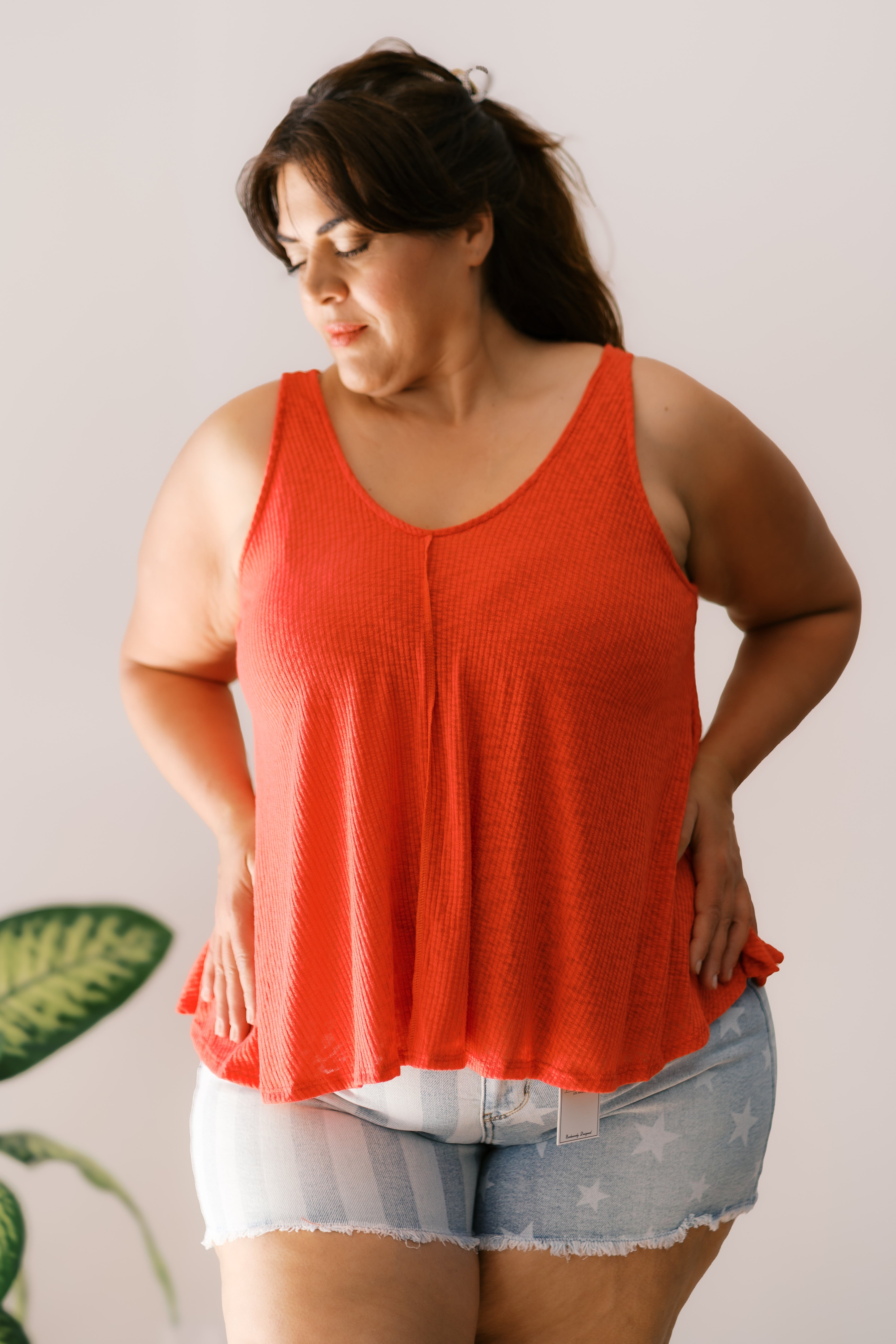 Plus size woman wearing sleeveless bright orange tank top and light denim shorts standing indoors near plant