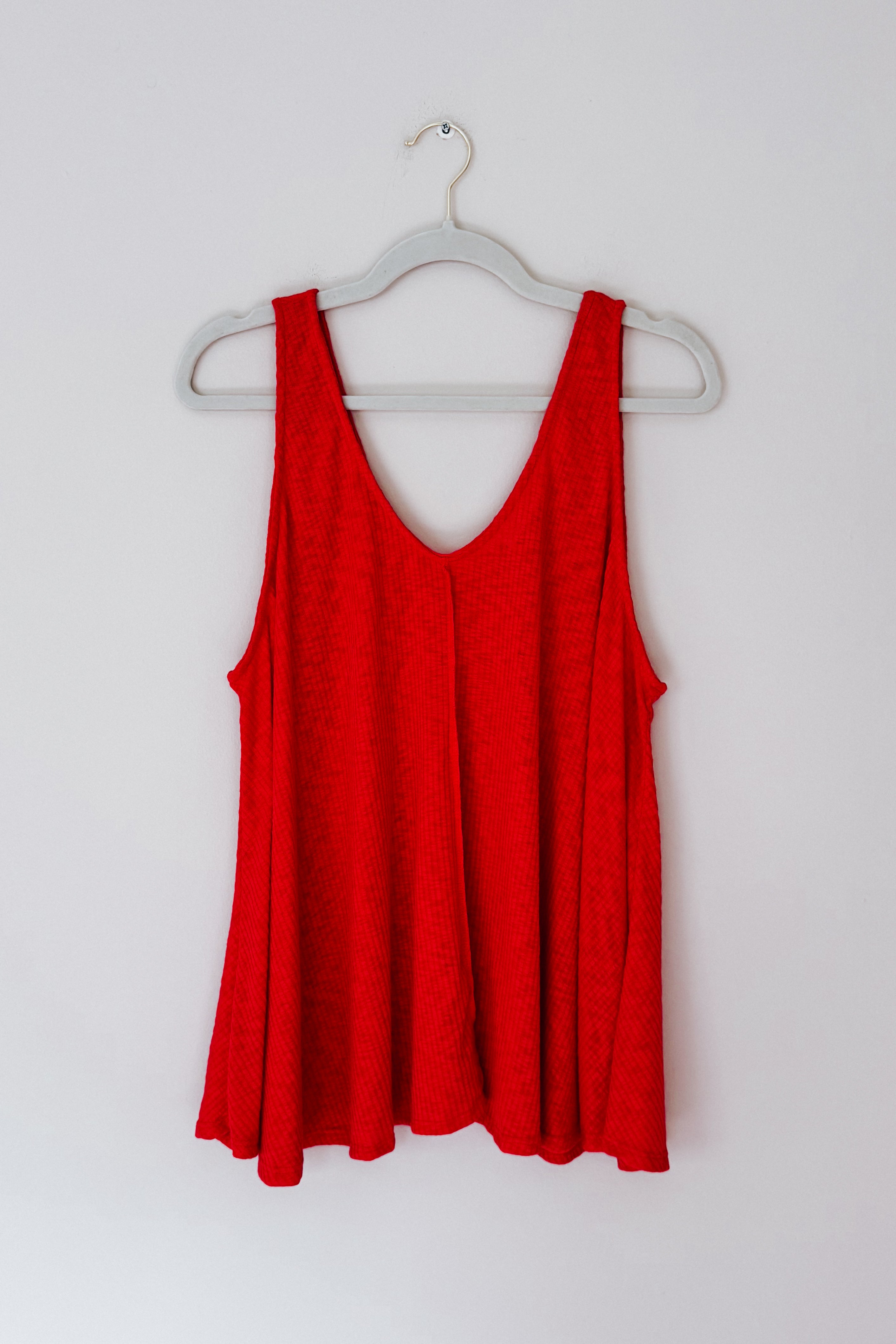 Bright red sleeveless tank top with textured fabric hanging on white plastic hanger against plain wall background