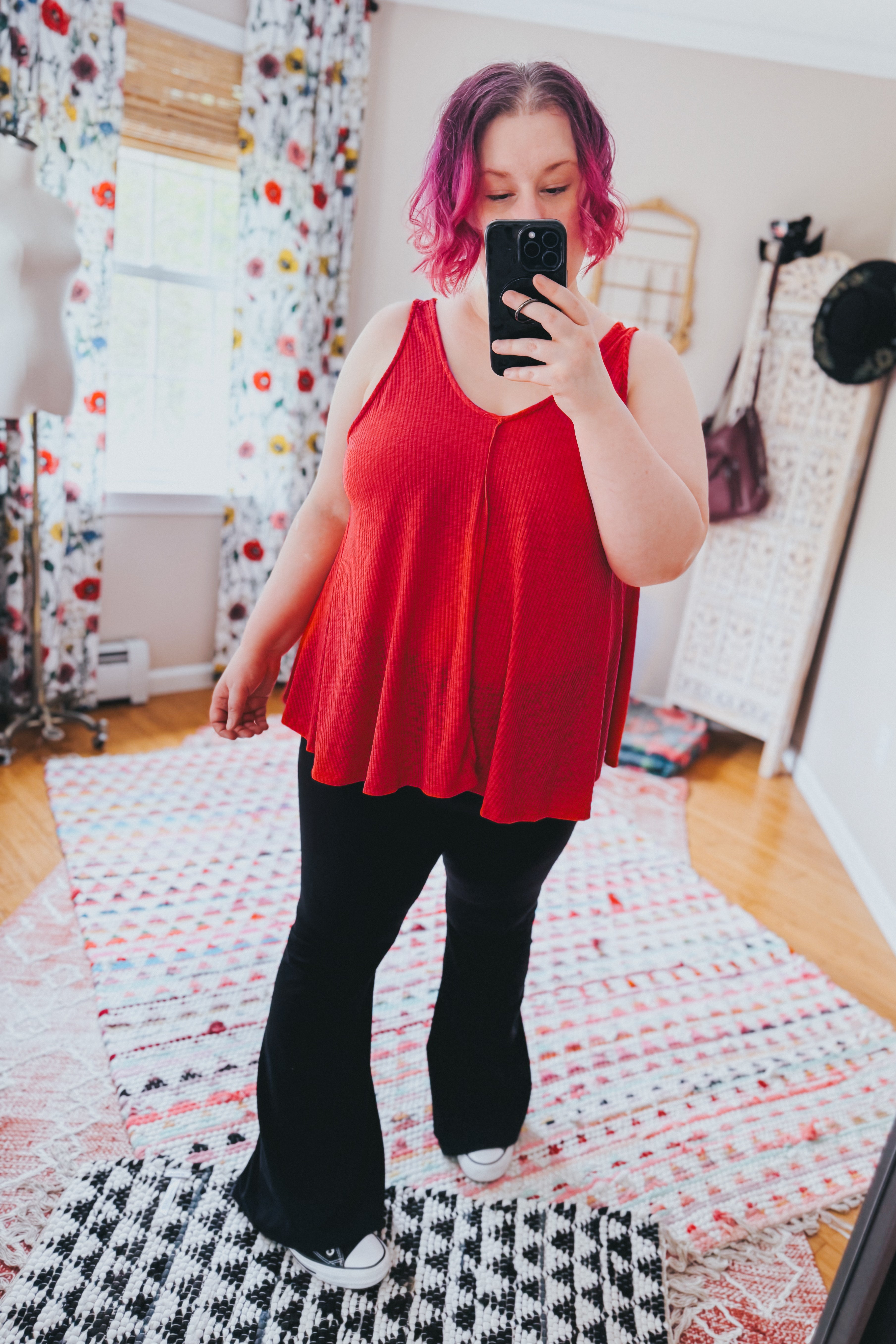 Plus size woman with short pink hair wearing a loose red sleeveless tank top and black flared pants indoors taking a phone selfie