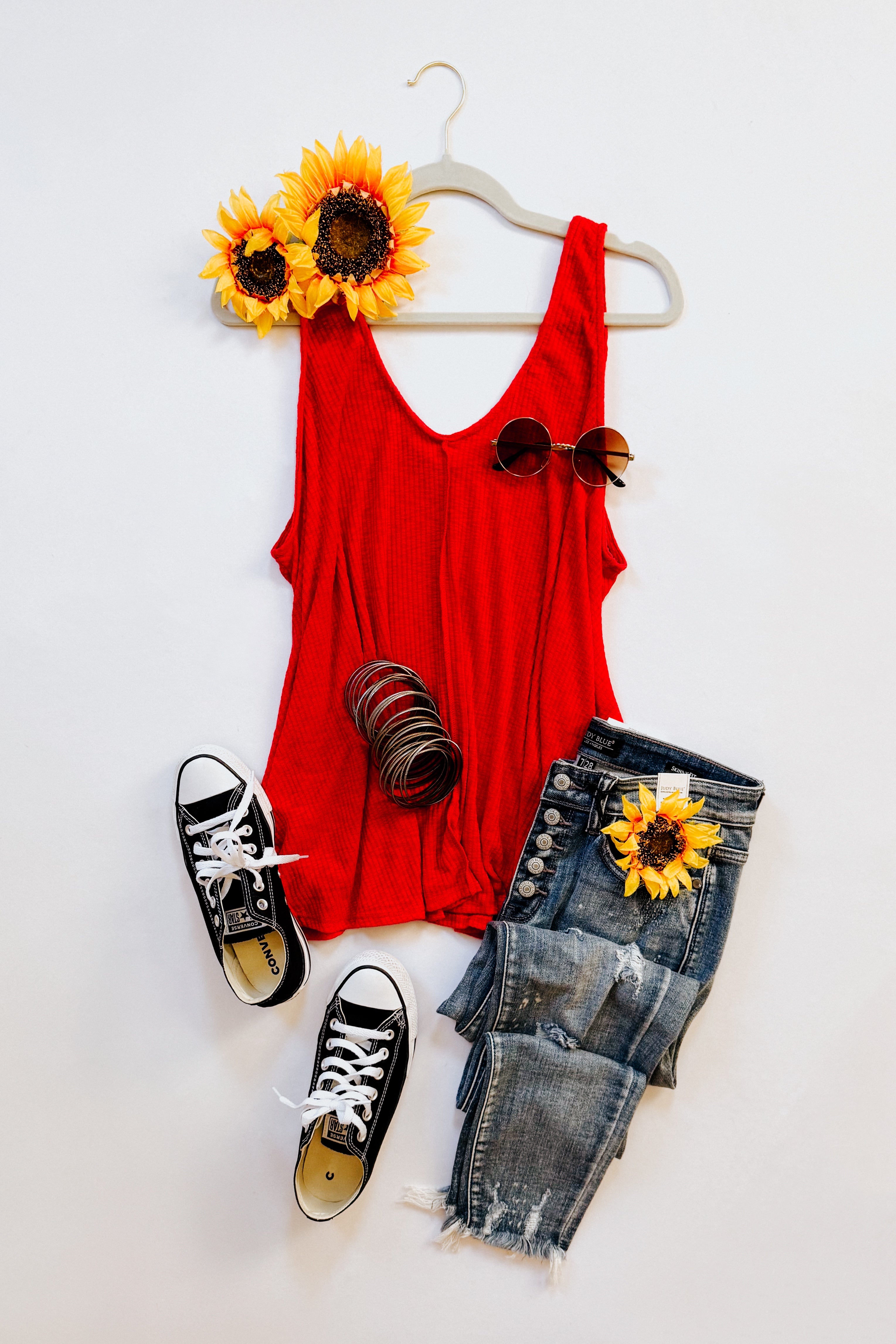 Bright red sleeveless tank top on a hanger paired with distressed button-fly jeans, black sneakers, sunflower, and sunglasses