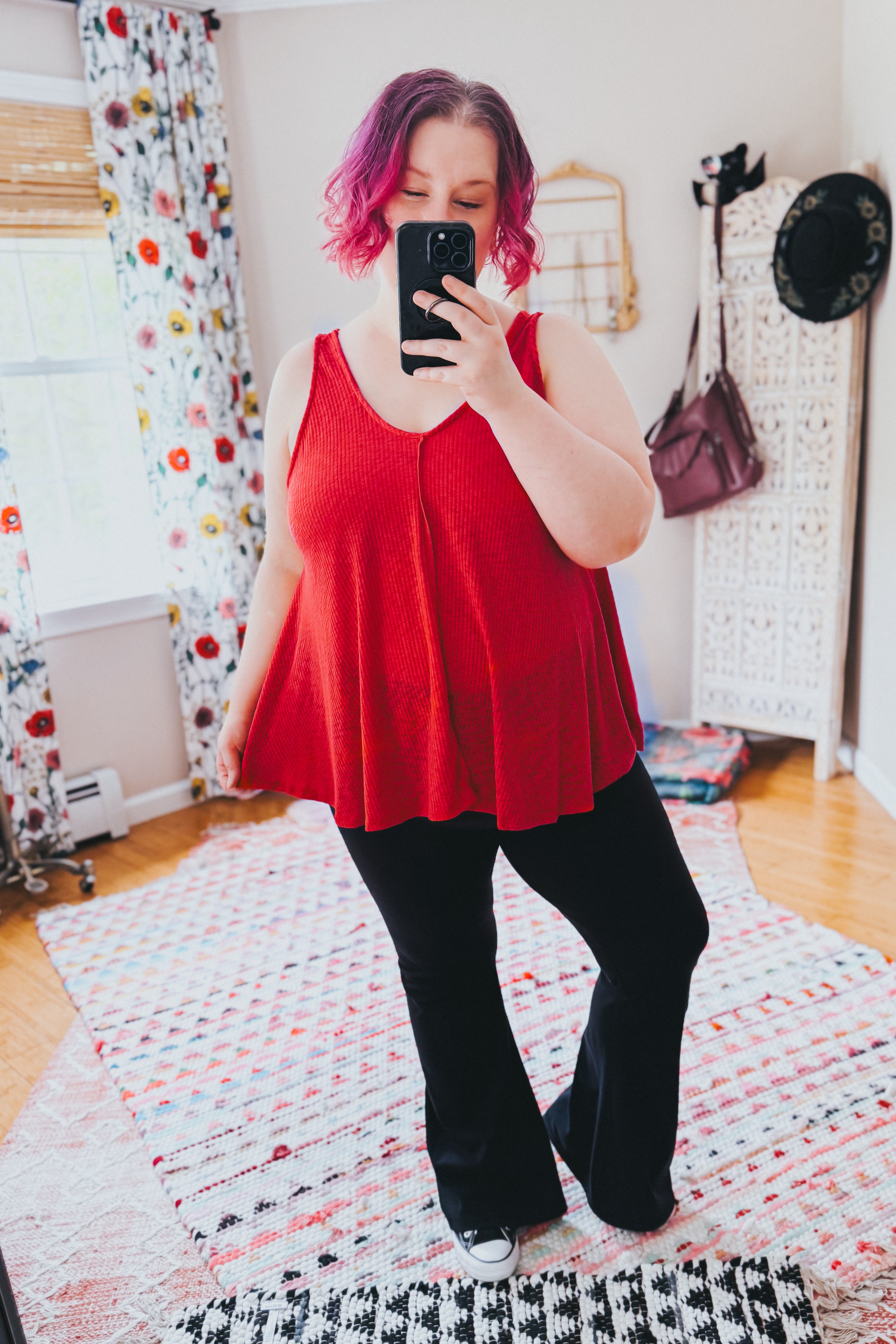 Plus size woman with short pink hair wearing a flowing red sleeveless tank top and black pants taking a selfie indoors
