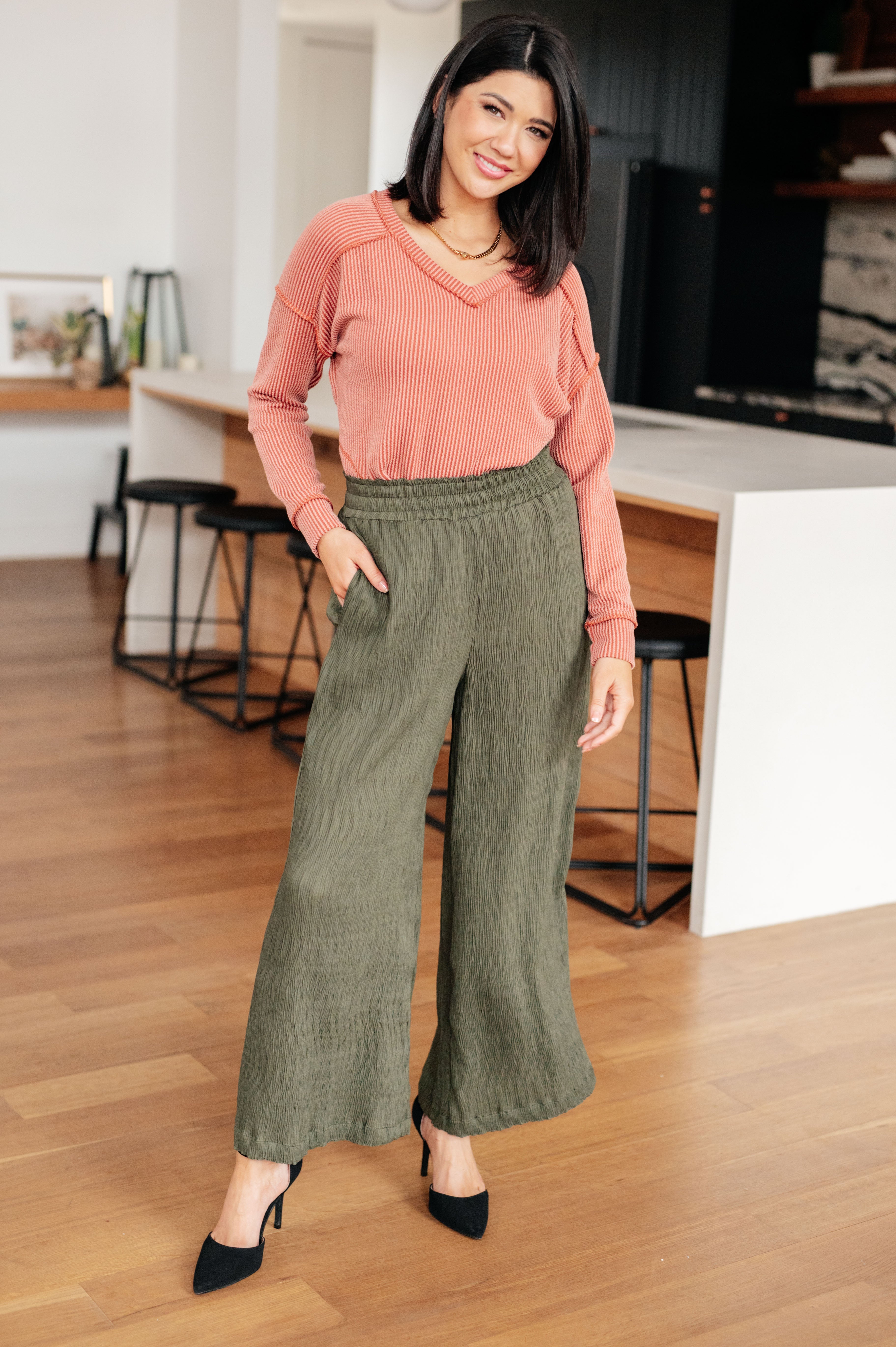 Smiling woman standing indoors wearing rust First and Foremost Rib Knit Top with V-neck and long sleeves paired with olive wide-leg pants and black heels in modern space