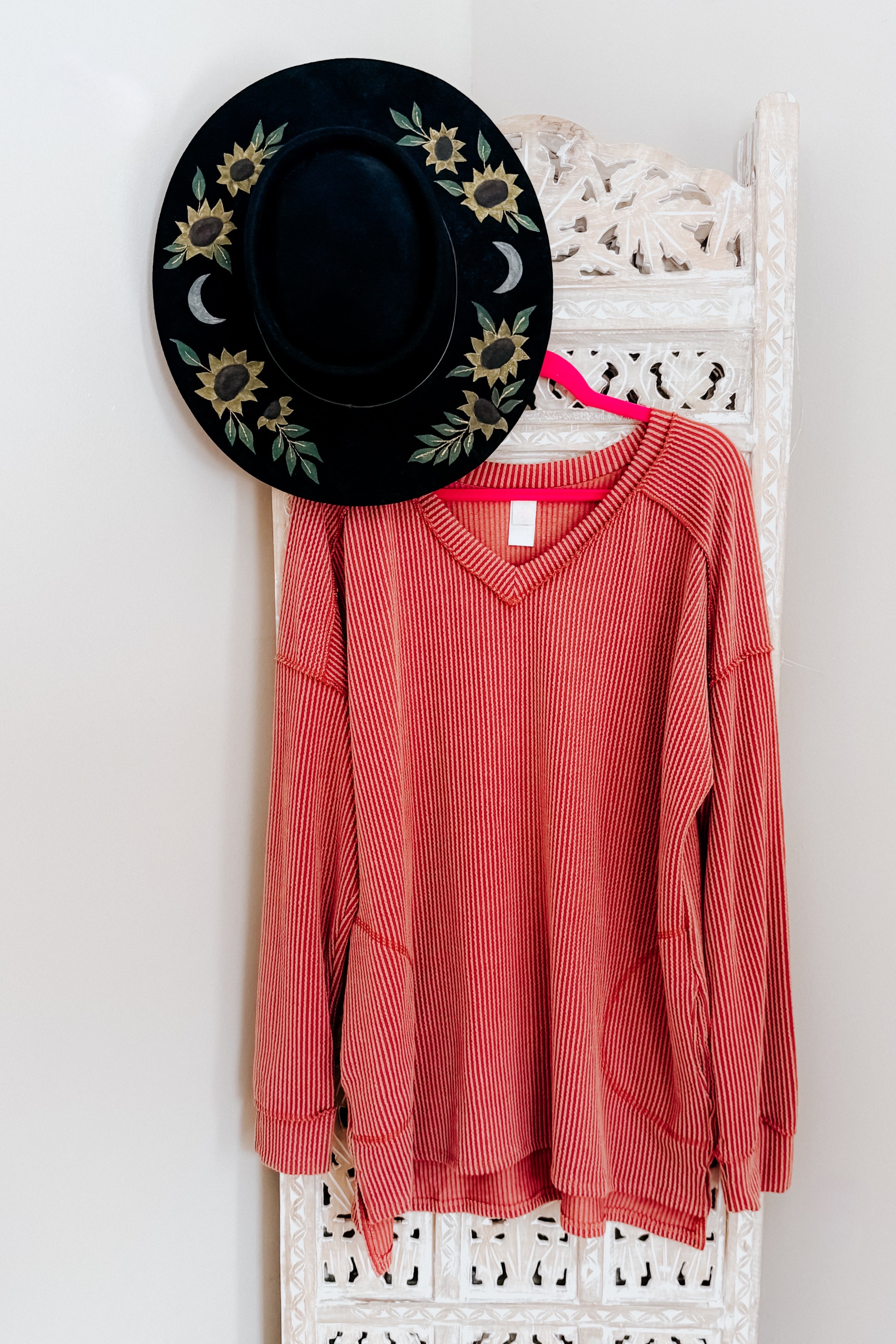 Rust ribbed V-neck First and Foremost Rib Knit Top hanging on bright pink hanger next to black wide brim hat with sunflower and moon embroidery on carved white panel