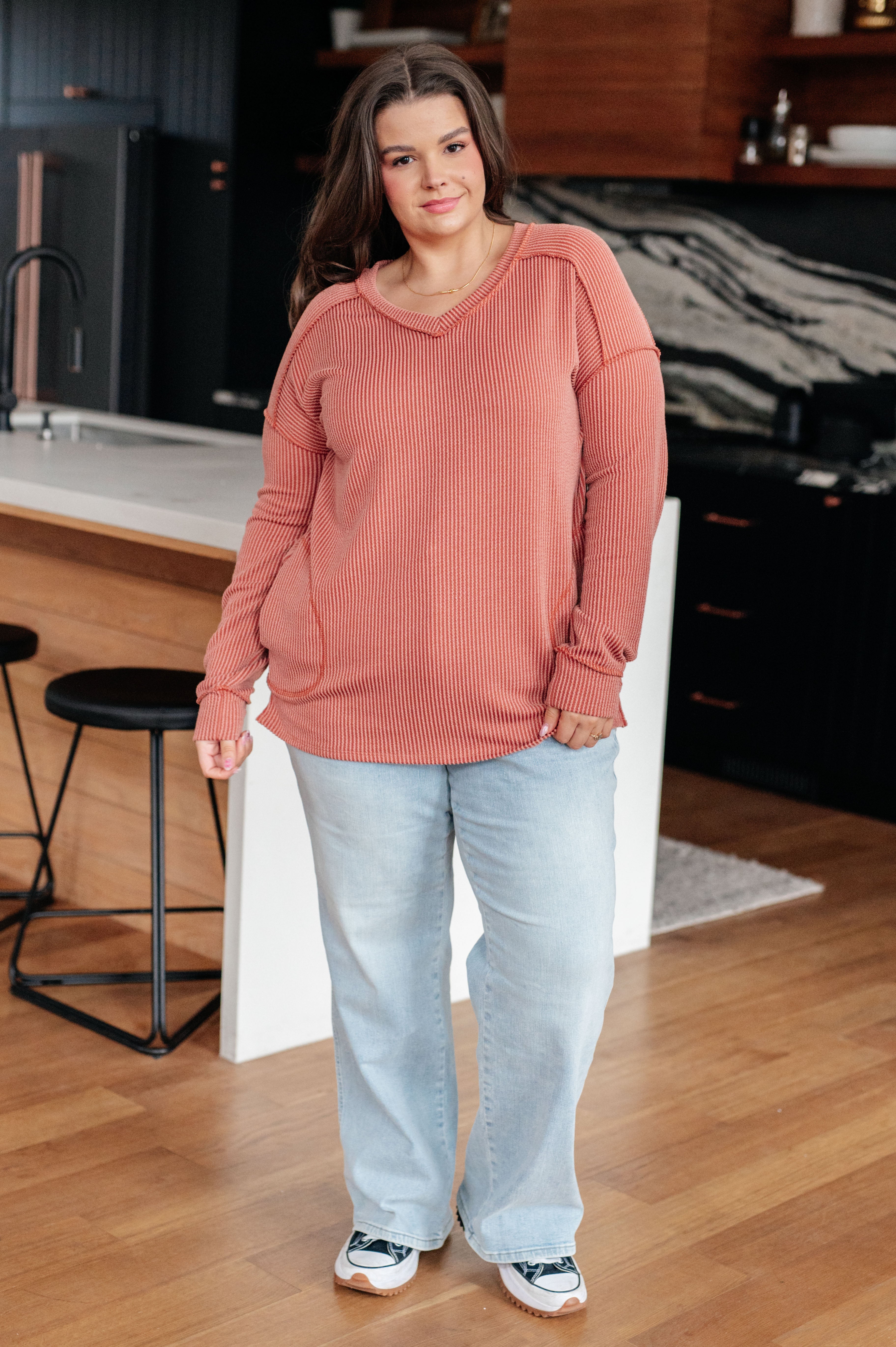 Woman wearing rust First and Foremost Rib Knit Top with relaxed fit and V-neck paired with light blue jeans and sneakers in stylish kitchen setting