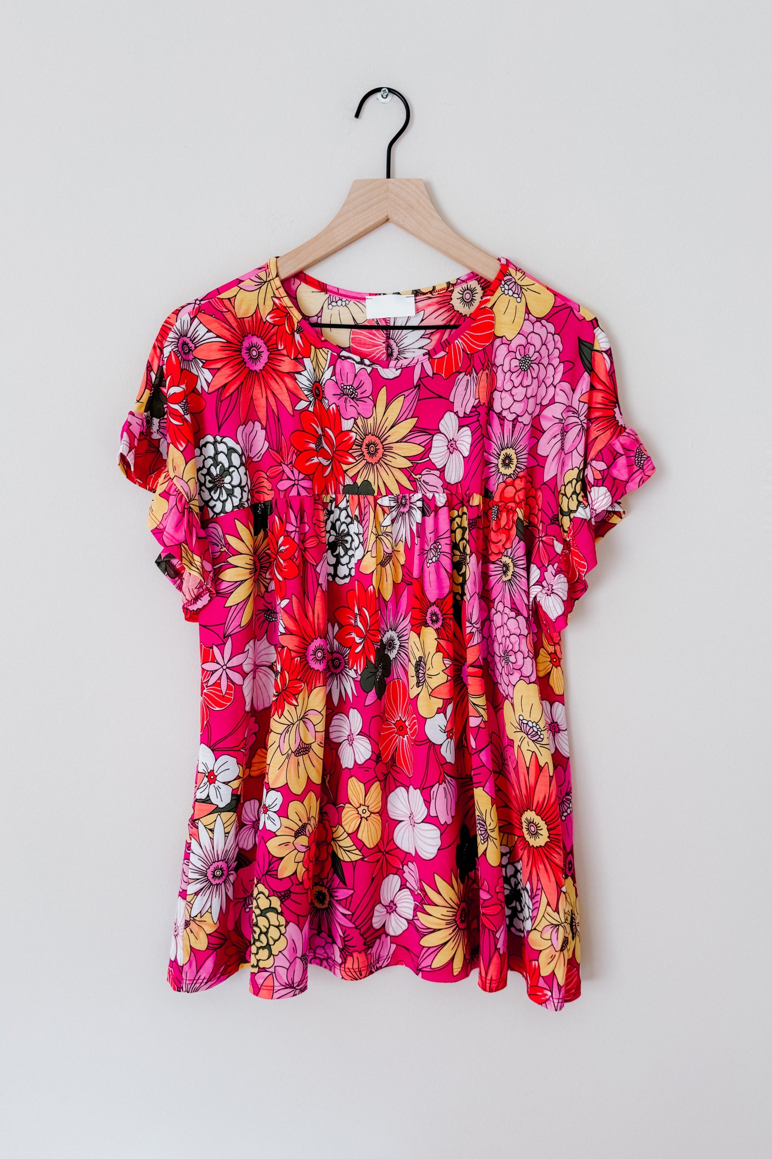 Flit About Floral Top in Pink with vibrant multicolor floral print and ruffled short sleeves displayed on wooden hanger against white background