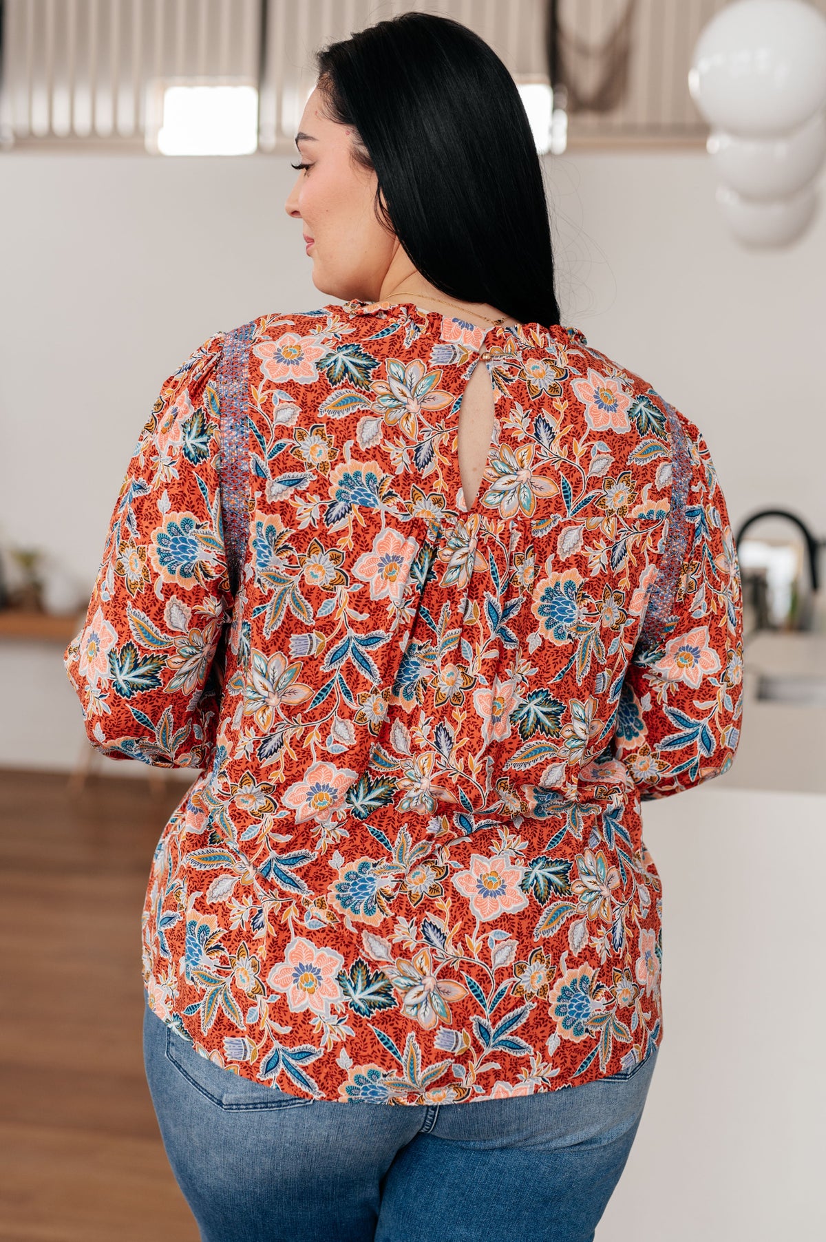 Back view of a woman wearing a Rust floral blouse with a ruffled neckline and blue jeans in a warmly lit home interior