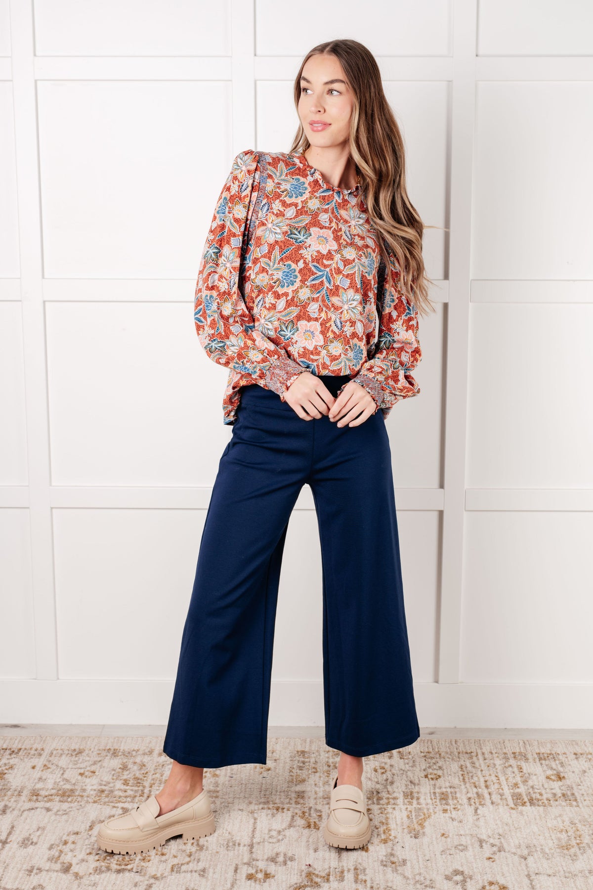 Woman modeling a rust floral ruffle neck blouse with navy wide leg pants and loafers in a bright paneled room