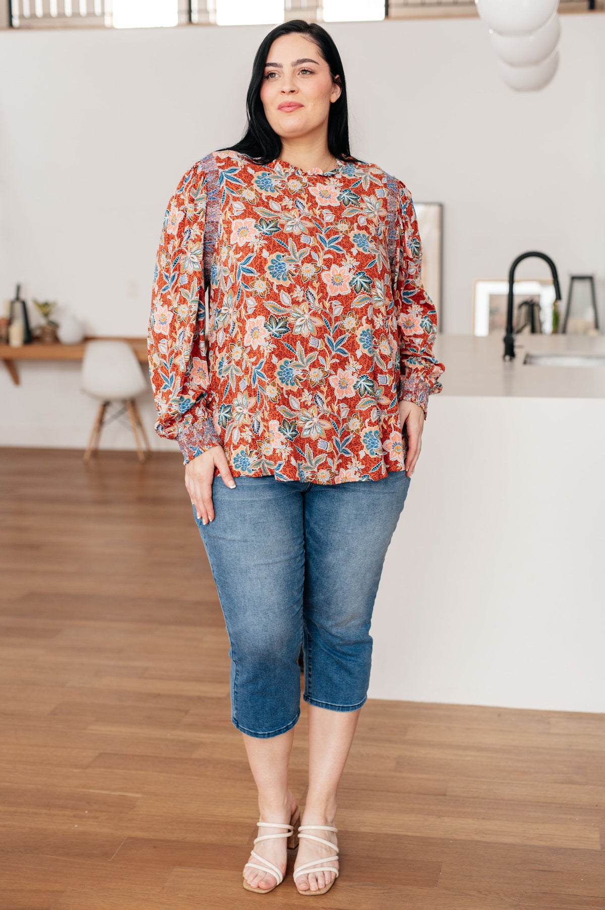 Woman modeling a rust floral blouse with a ruffled neckline and long sleeves with cropped jeans and strappy sandals in a bright home interior