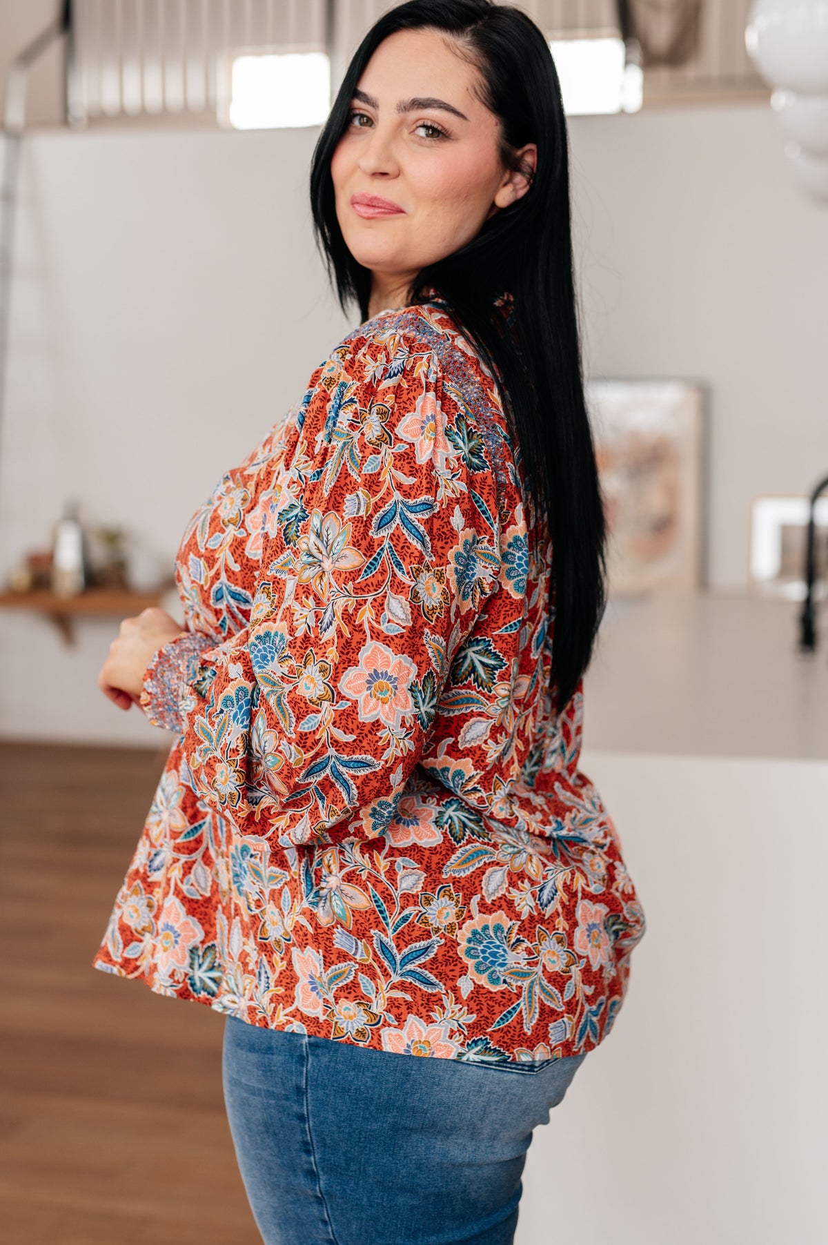 Woman modeling a rust floral ruffle neck blouse with long sleeves and blue jeans in a bright modern home interior