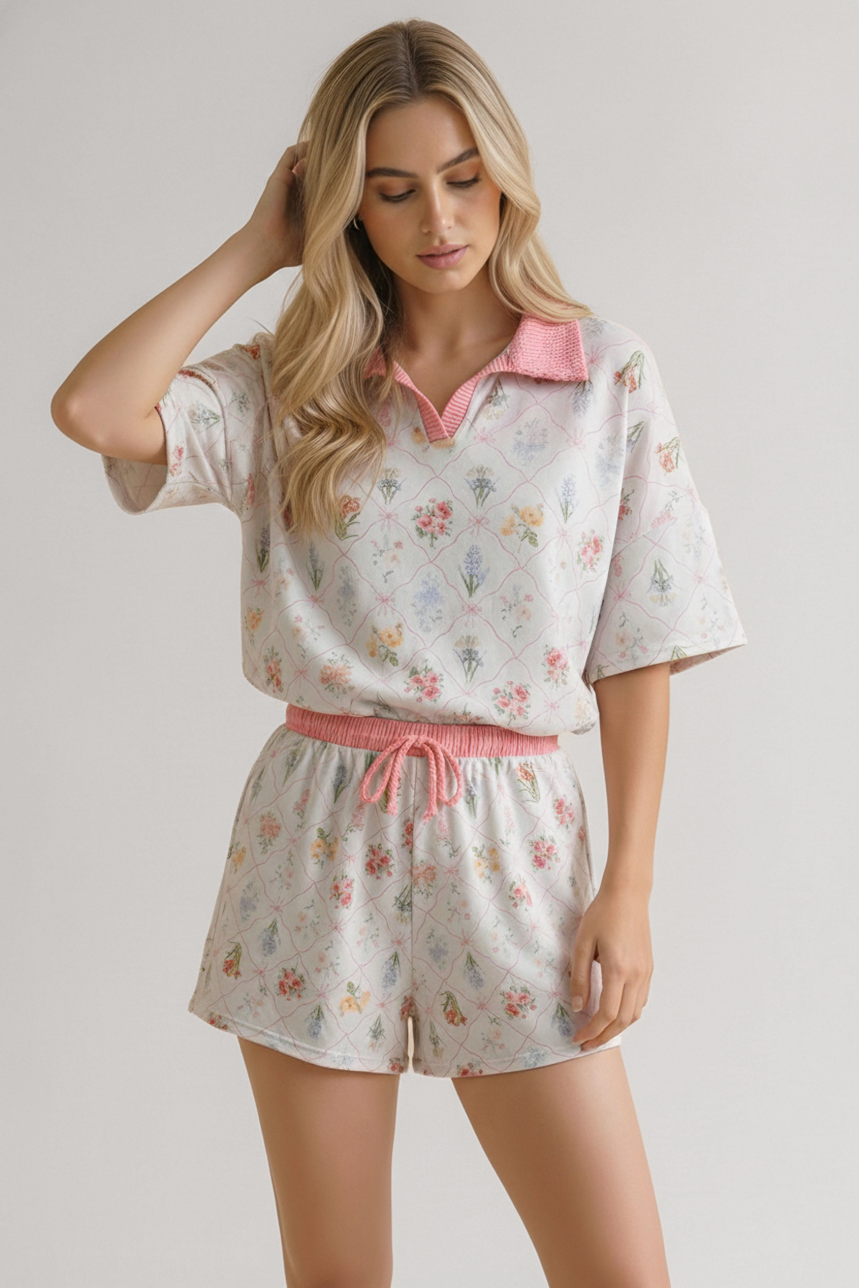 Woman wearing ivory floral garden short set with pink collar and waistband, standing with one hand in hair
