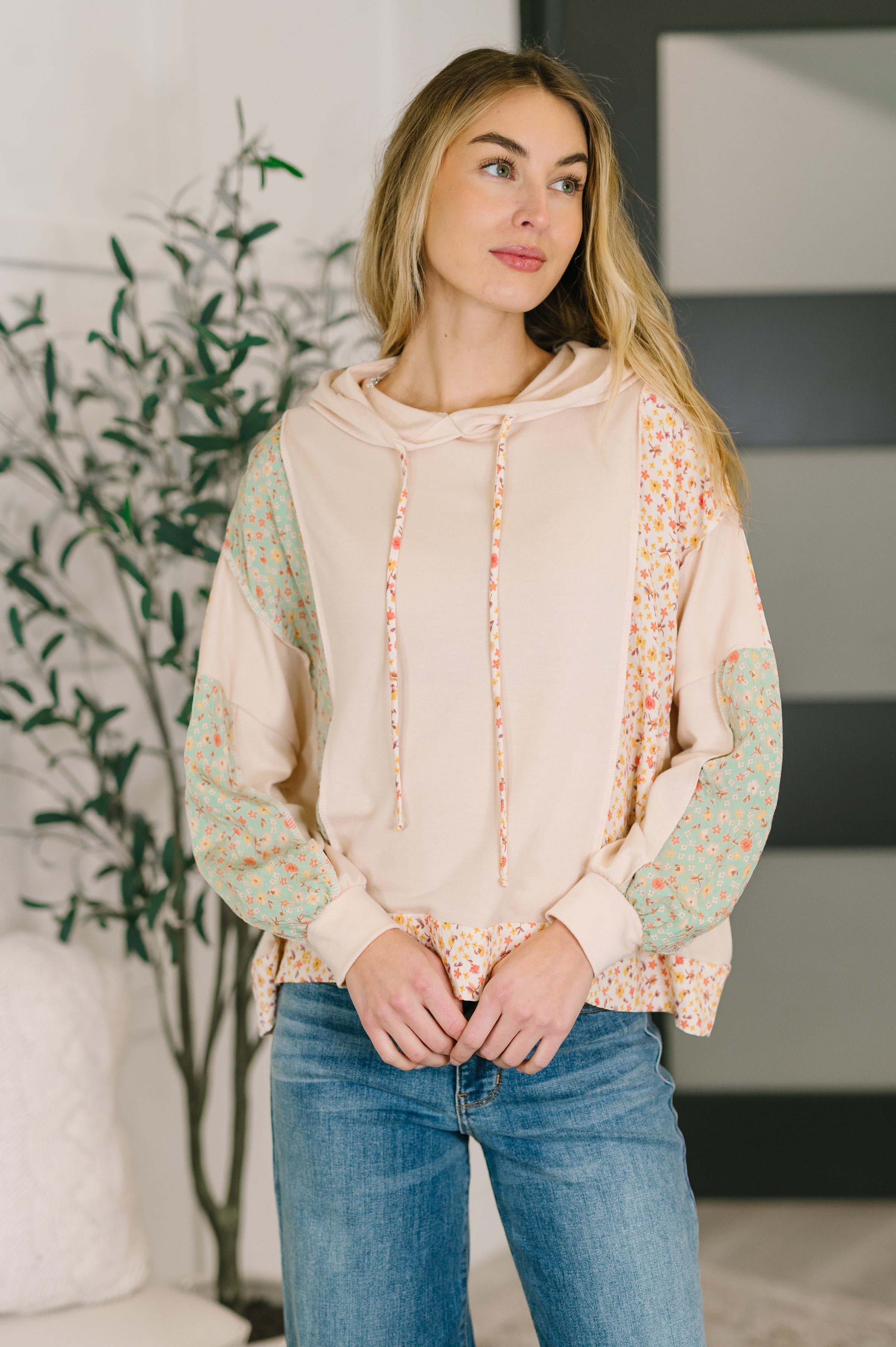 Woman indoors wearing cream Floral Patchwork Hooded Pullover with green and orange floral sleeve panels paired with blue jeans