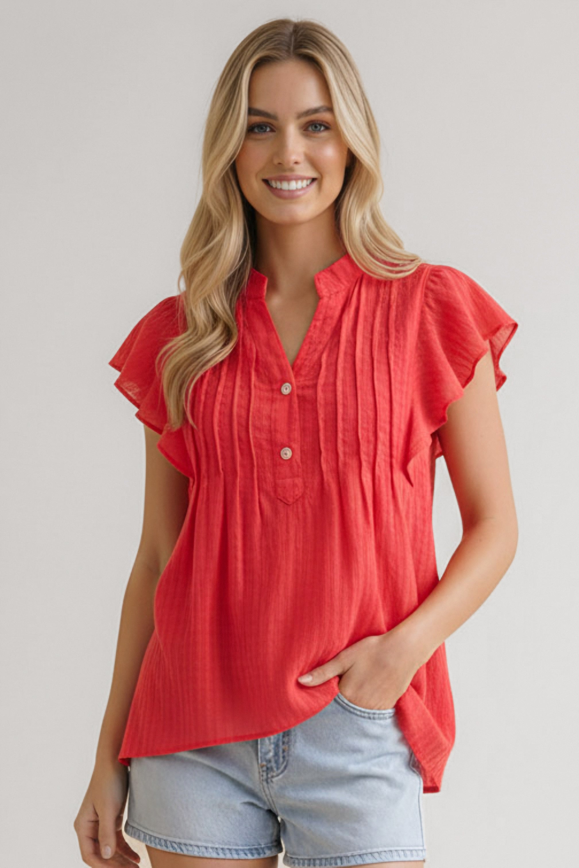Woman wearing coral flutter sleeve top with pintuck details and light denim shorts smiling indoors