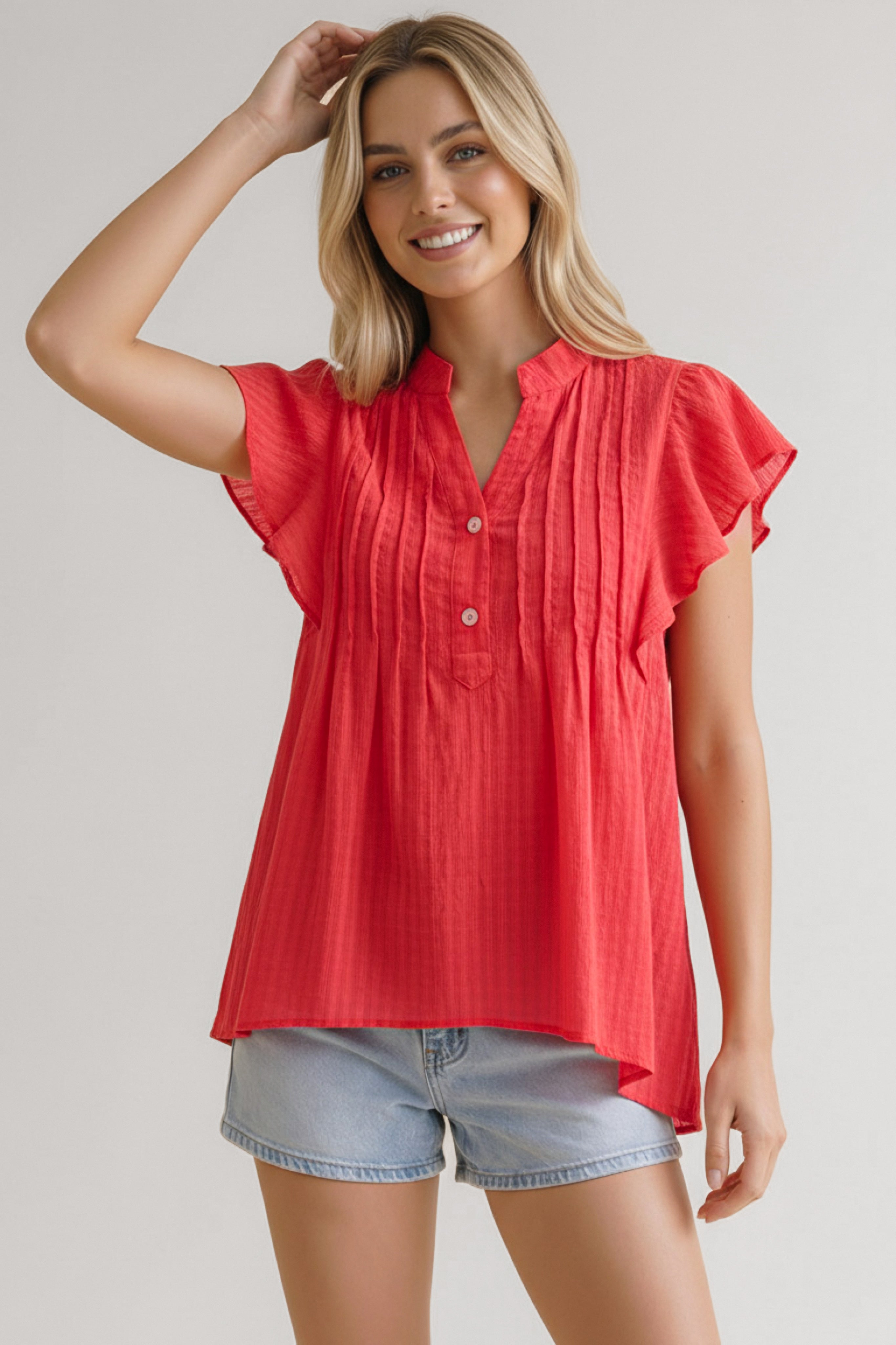 Smiling woman wearing coral flutter sleeve top with pintuck details paired with light denim shorts against plain background