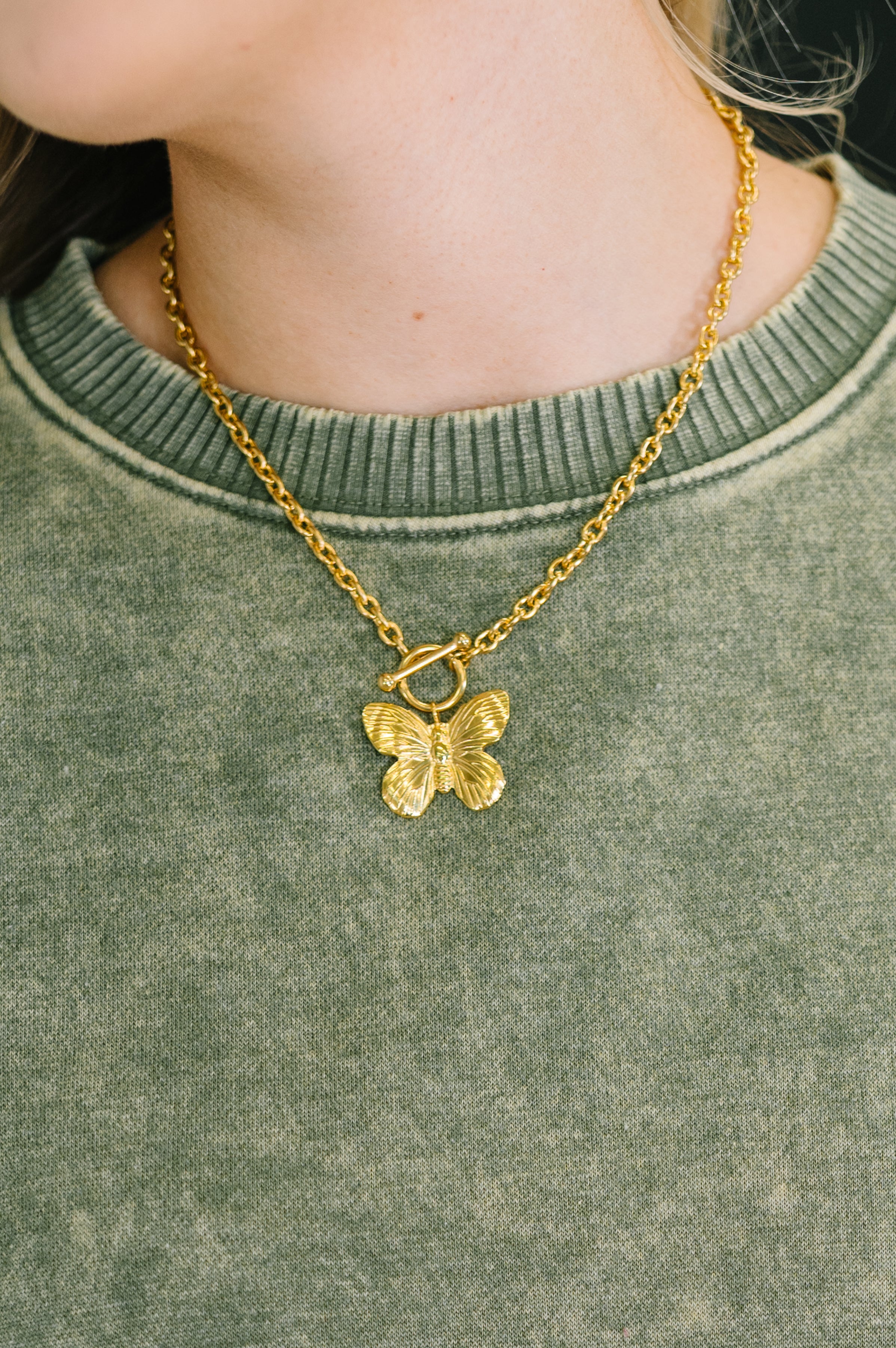 Flutter & Shine Butterfly Pendant Necklace • Gold worn on model with green textured top, showing detailed butterfly charm and chain links